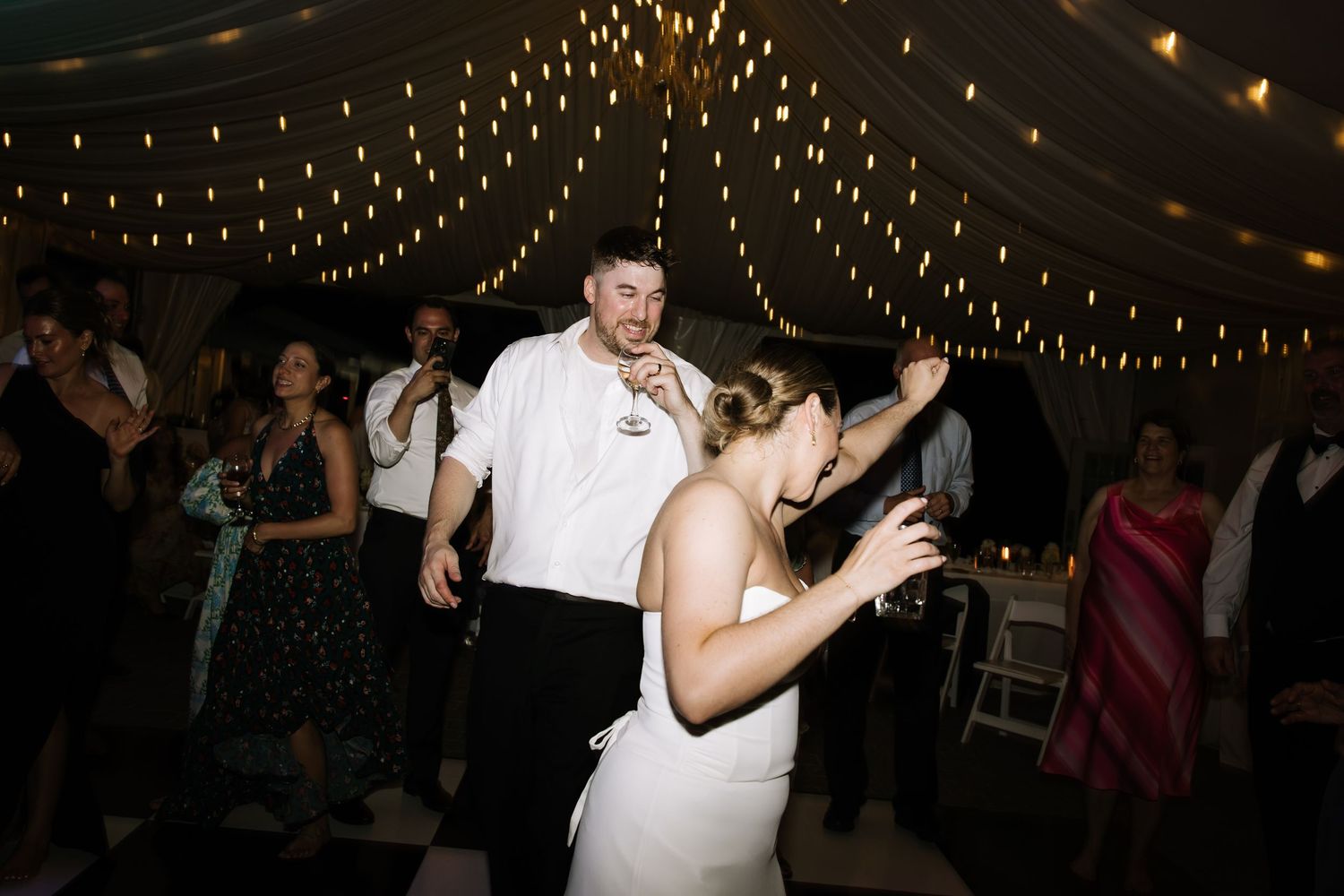 Bride and groom dancing joyfully at wedding reception under twinkling fairy lights with guests celebrating.