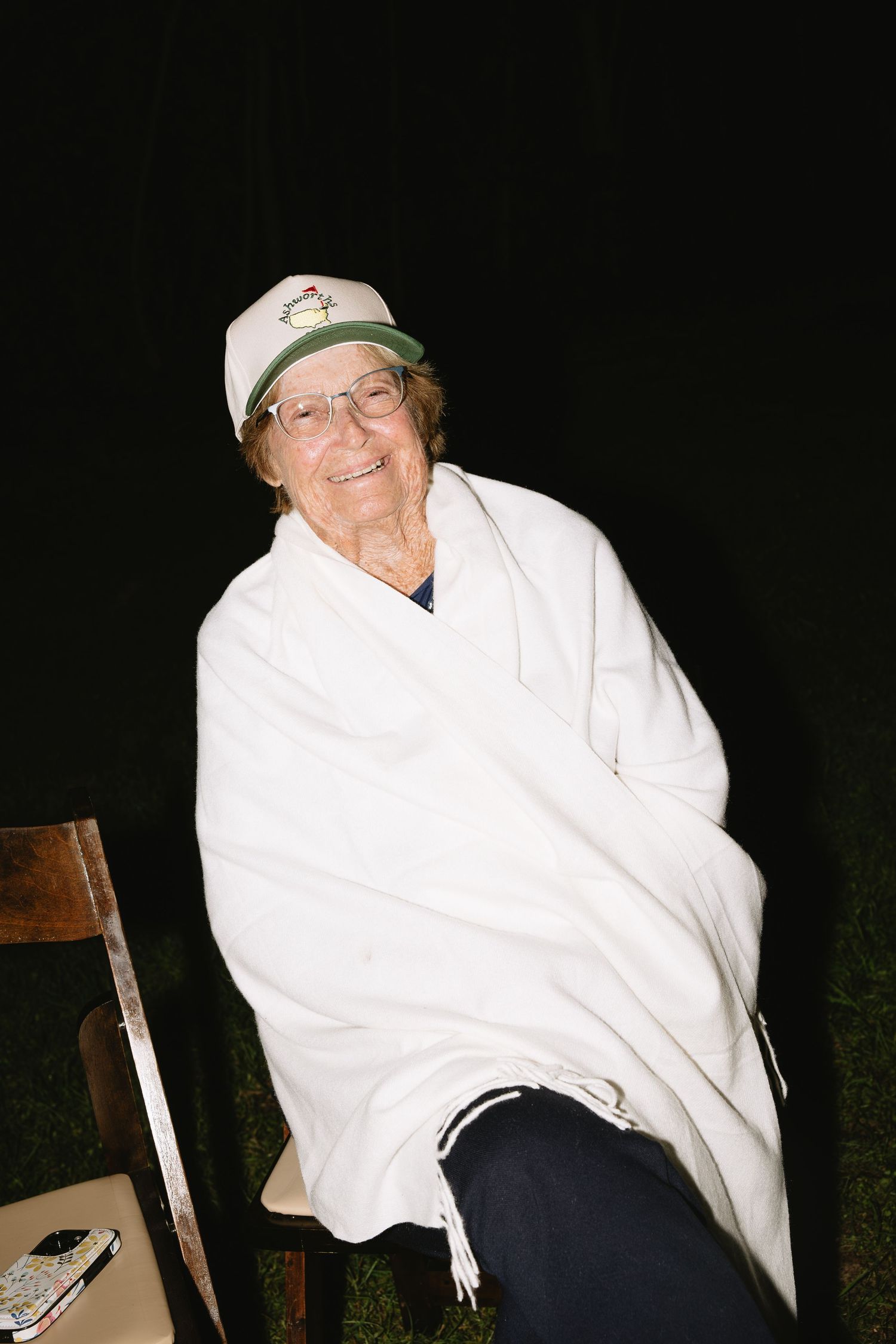 Elderly woman smiling wrapped in white blanket wearing green cap and glasses sitting on wooden chair outdoors at night.