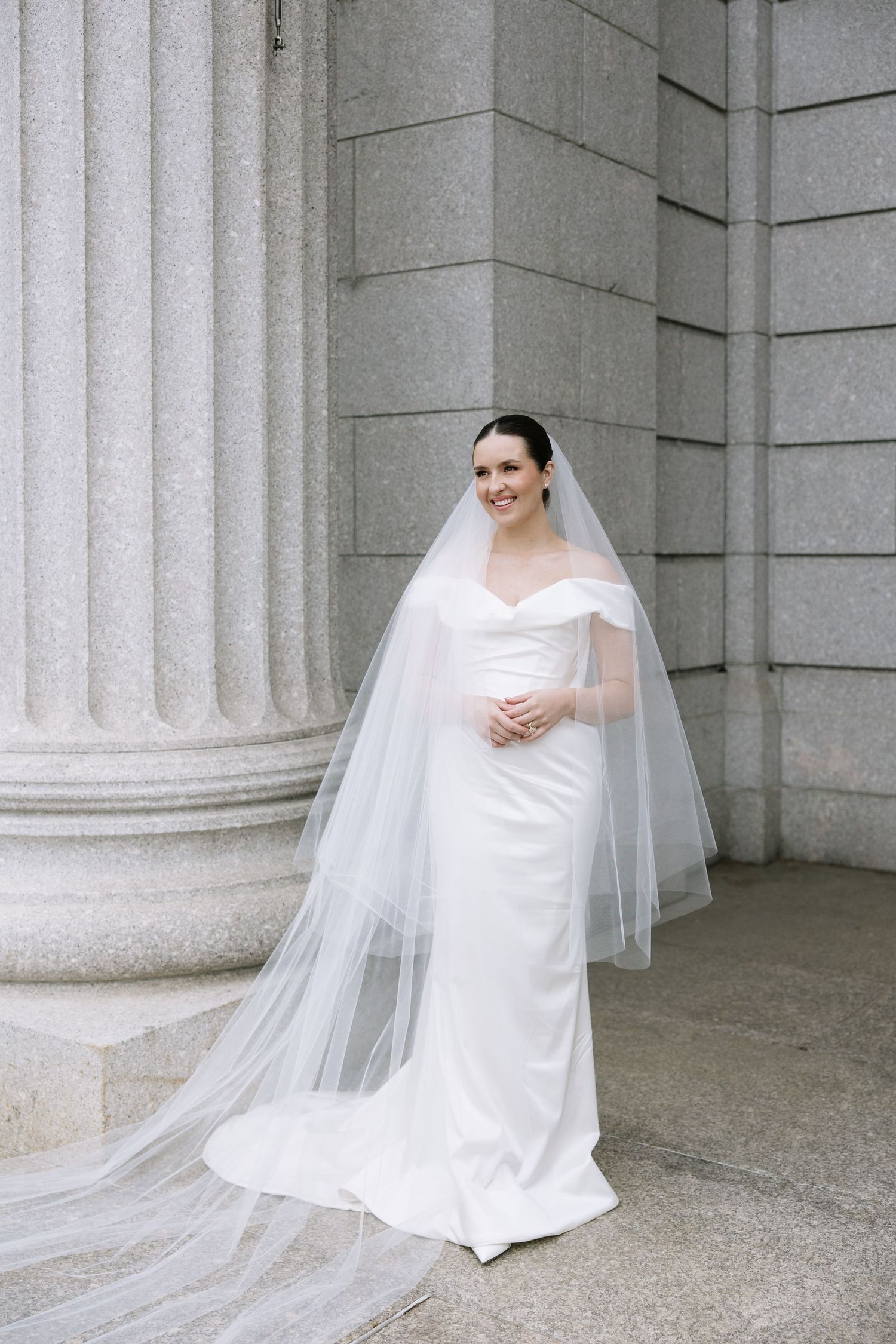 Bride in off-shoulder white gown and cathedral veil poses gracefully beside grand stone columns.