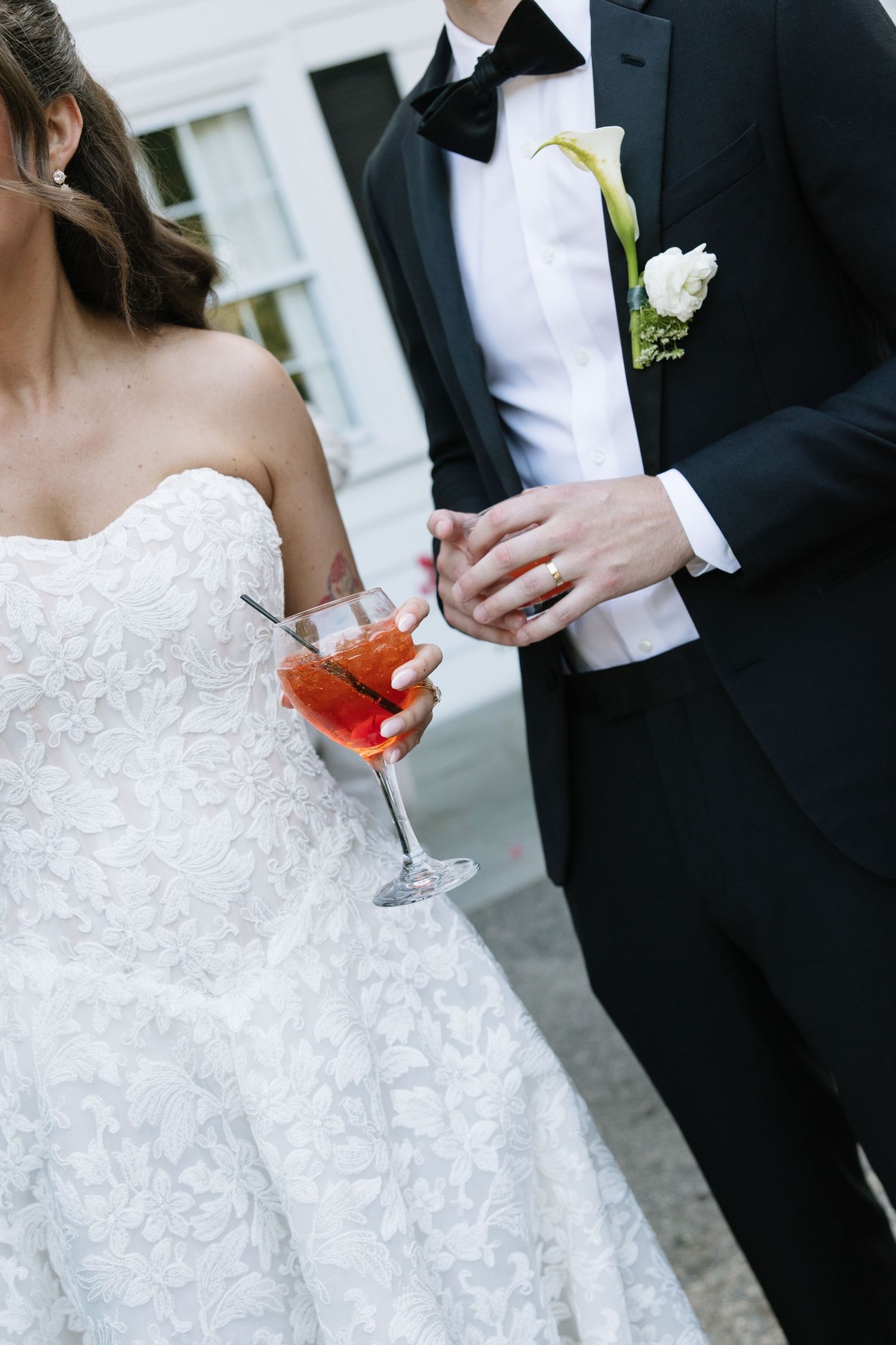 Bride in lace gown holding a red cocktail standing beside groom in black tuxedo with floral boutonniere.