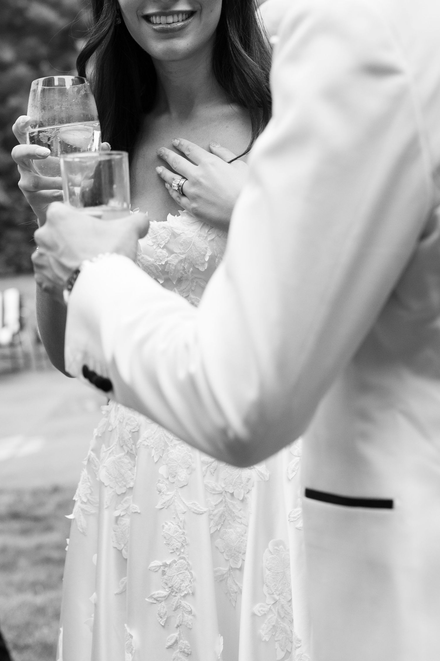 Black and white photo of smiling bride in lace gown holding champagne glass with groom in white tuxedo jacket.