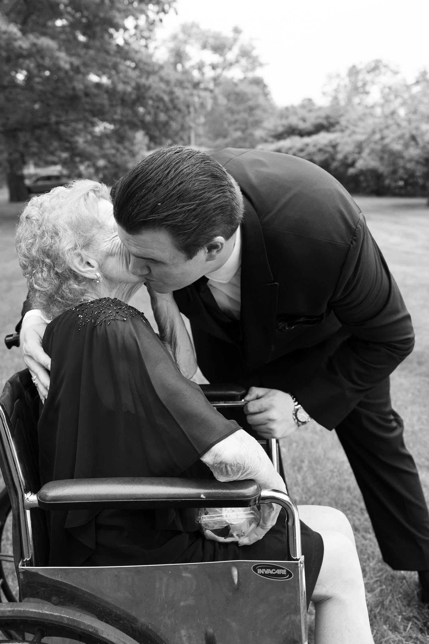 A man in a black suit kisses an elderly woman in a wheelchair outdoors in a black and white wedding photo.