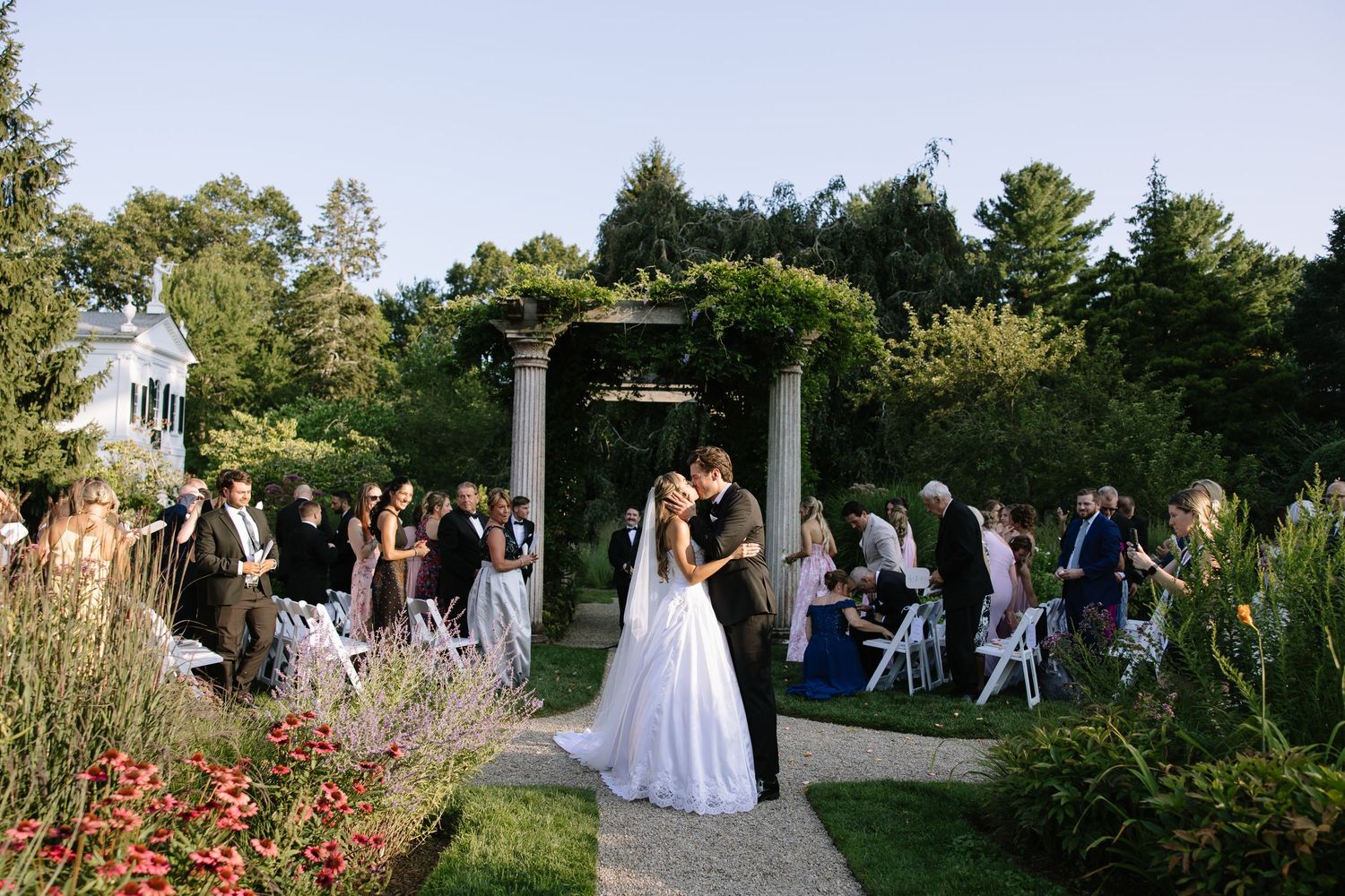 Wedding ceremony in a garden setting with stone columns and flowering plants.