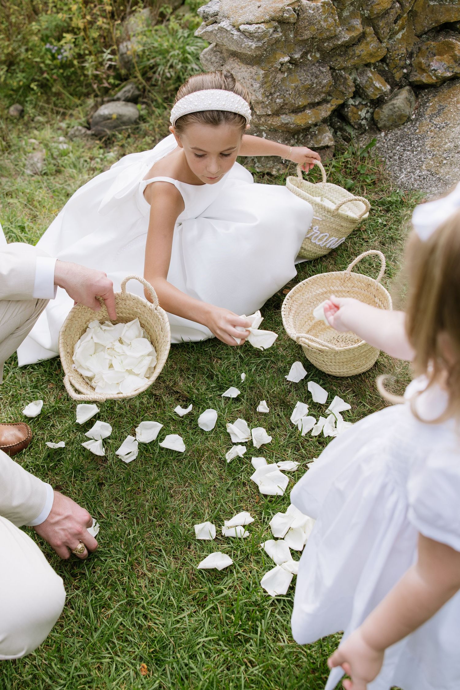 Young flower girls in white dresses scatter petals on grass during wedding ceremony.