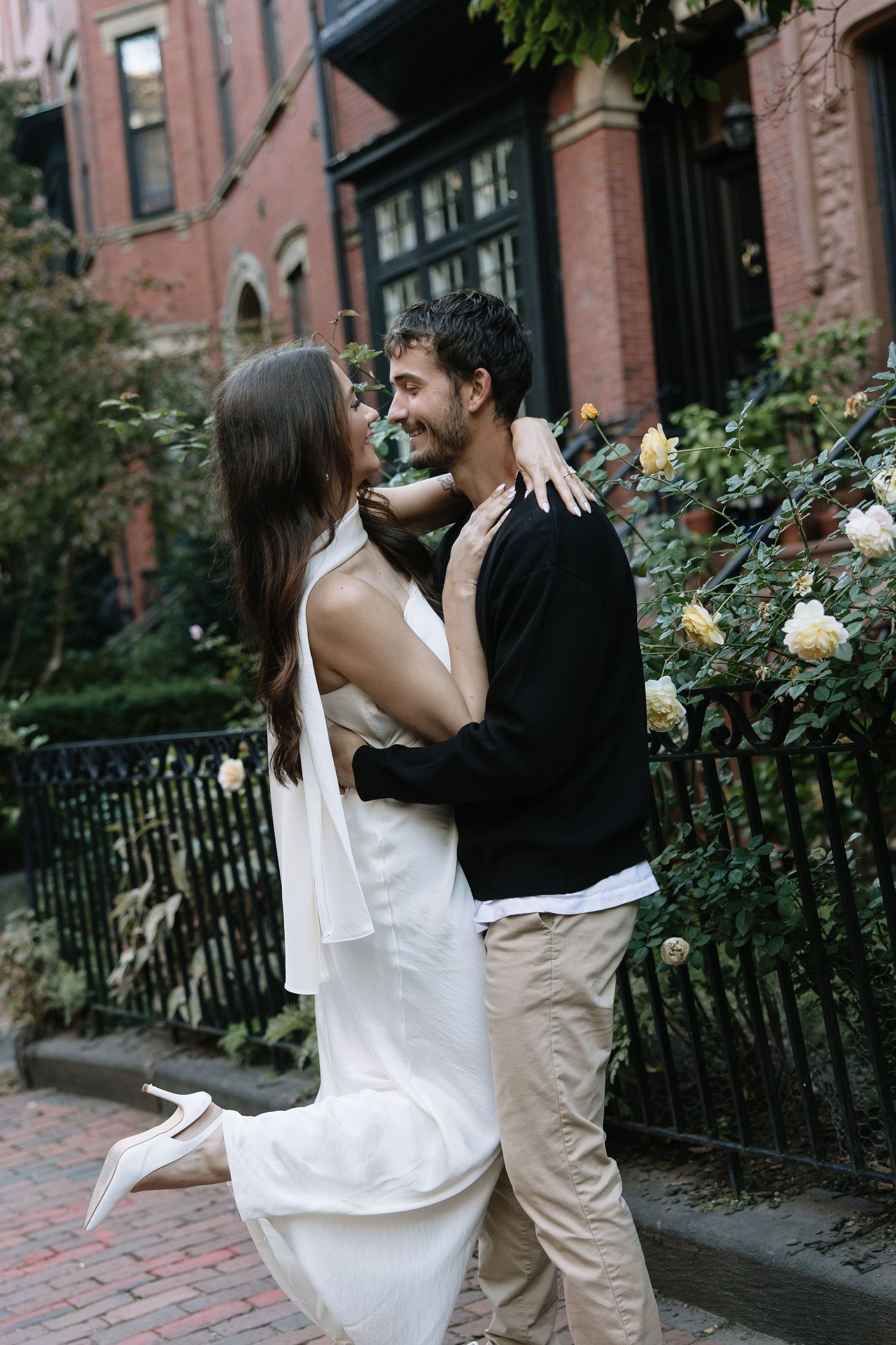 Couple posing by rose bushes outside historic brick buildings.