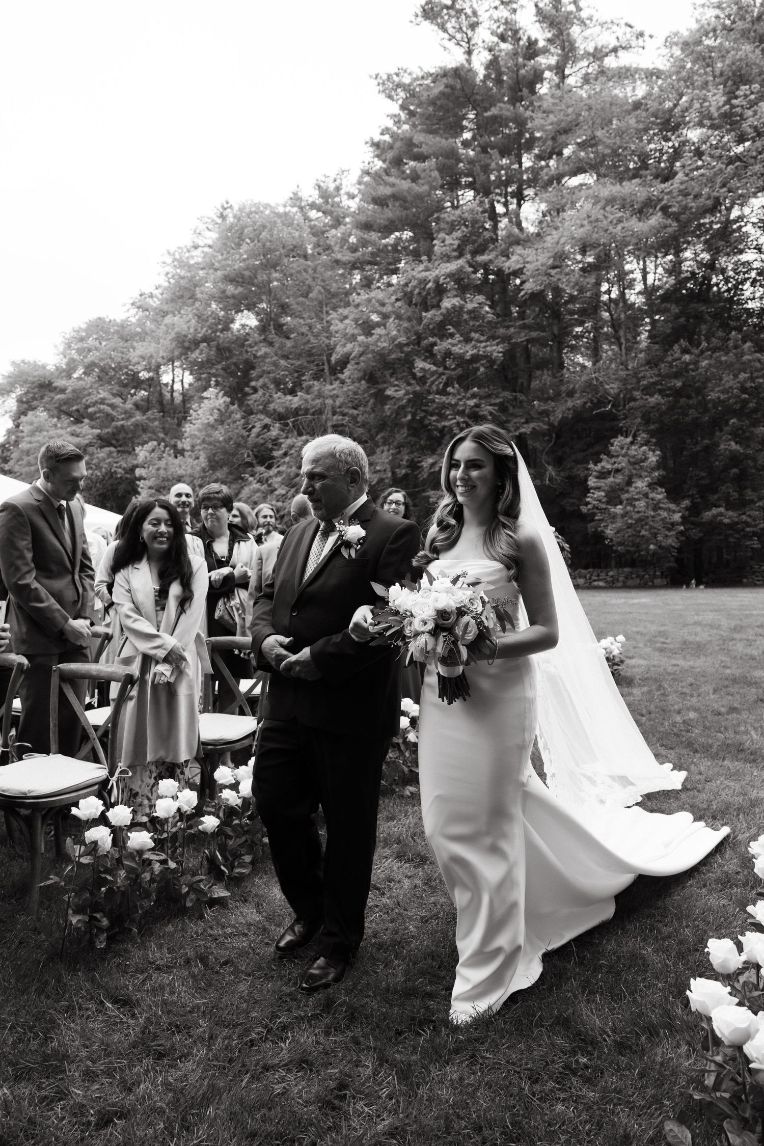 A bride in an elegant white gown walks down the aisle during an outdoor woodland ceremony.