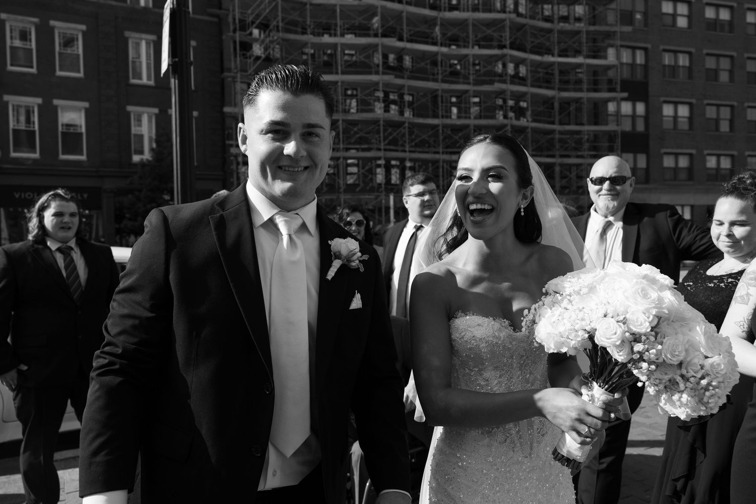 Wedding party poses for photos outside urban building with brick facade and decorative flowers.