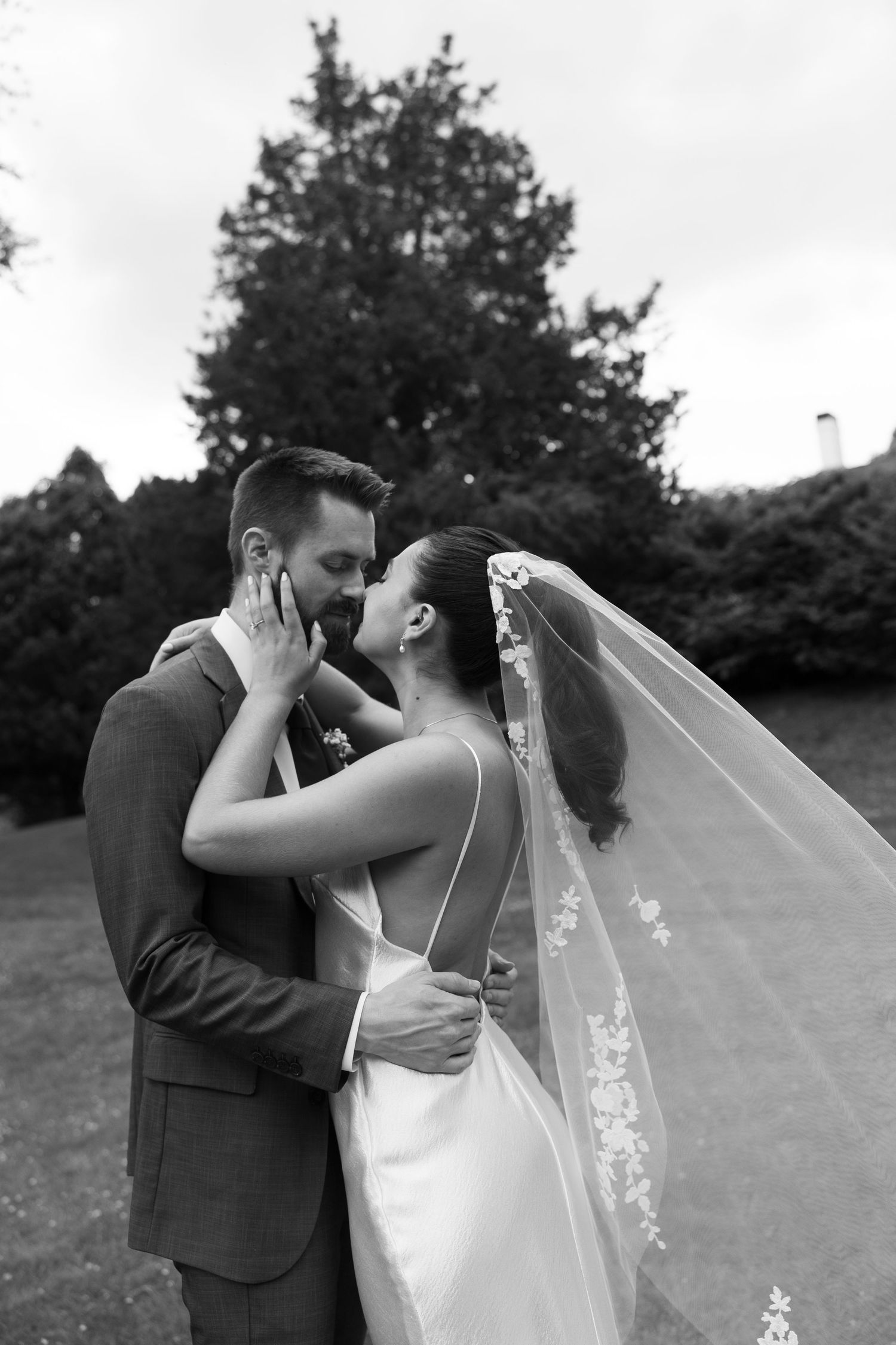 Black and white photo of a bride and groom sharing a romantic kiss outdoors, with her lace veil flowing in the breeze.