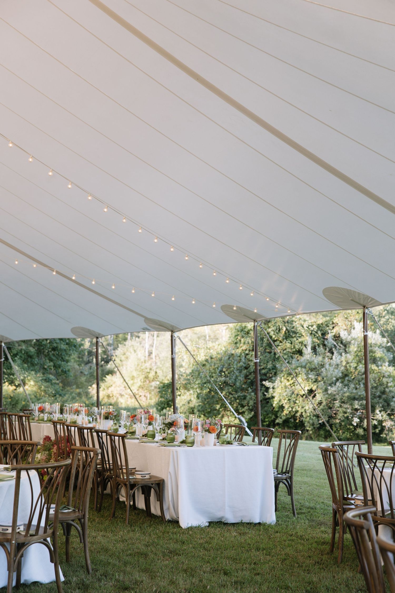 Elegant outdoor wedding reception under a white sailcloth tent with string lights, wooden chairs, and floral centerpieces on grass.