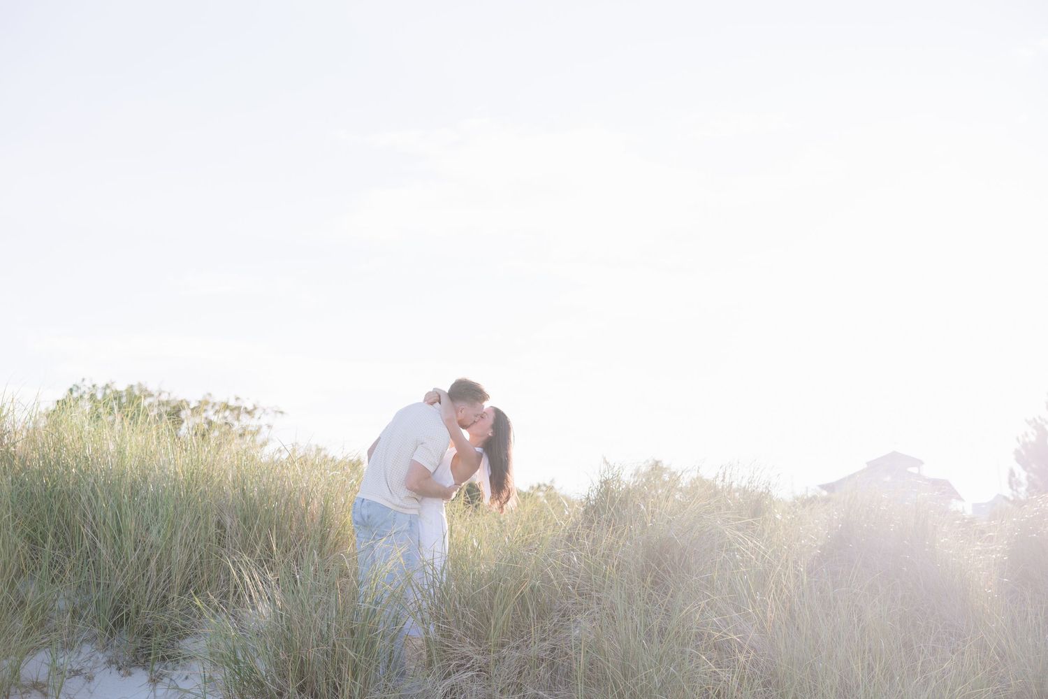 Couple kissing romantically on sandy beach dunes surrounded by tall grass in soft natural sunlight.