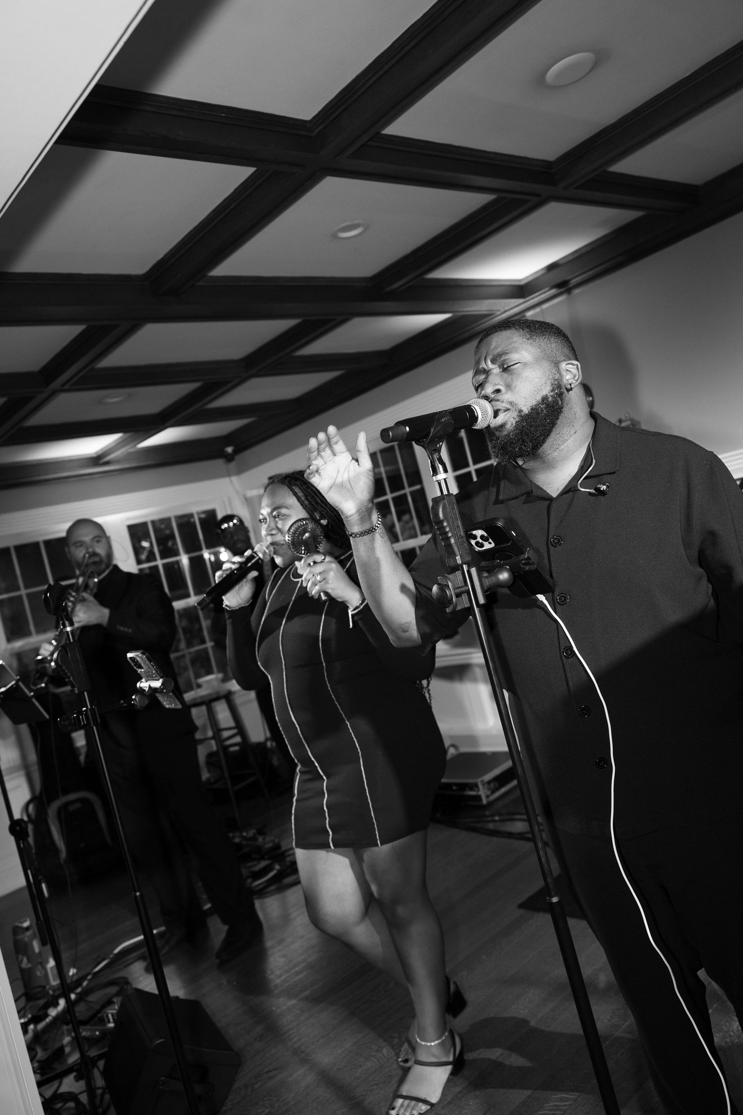 Black and white photo of a live band performing indoors, with a male vocalist singing passionately at a microphone stand.
