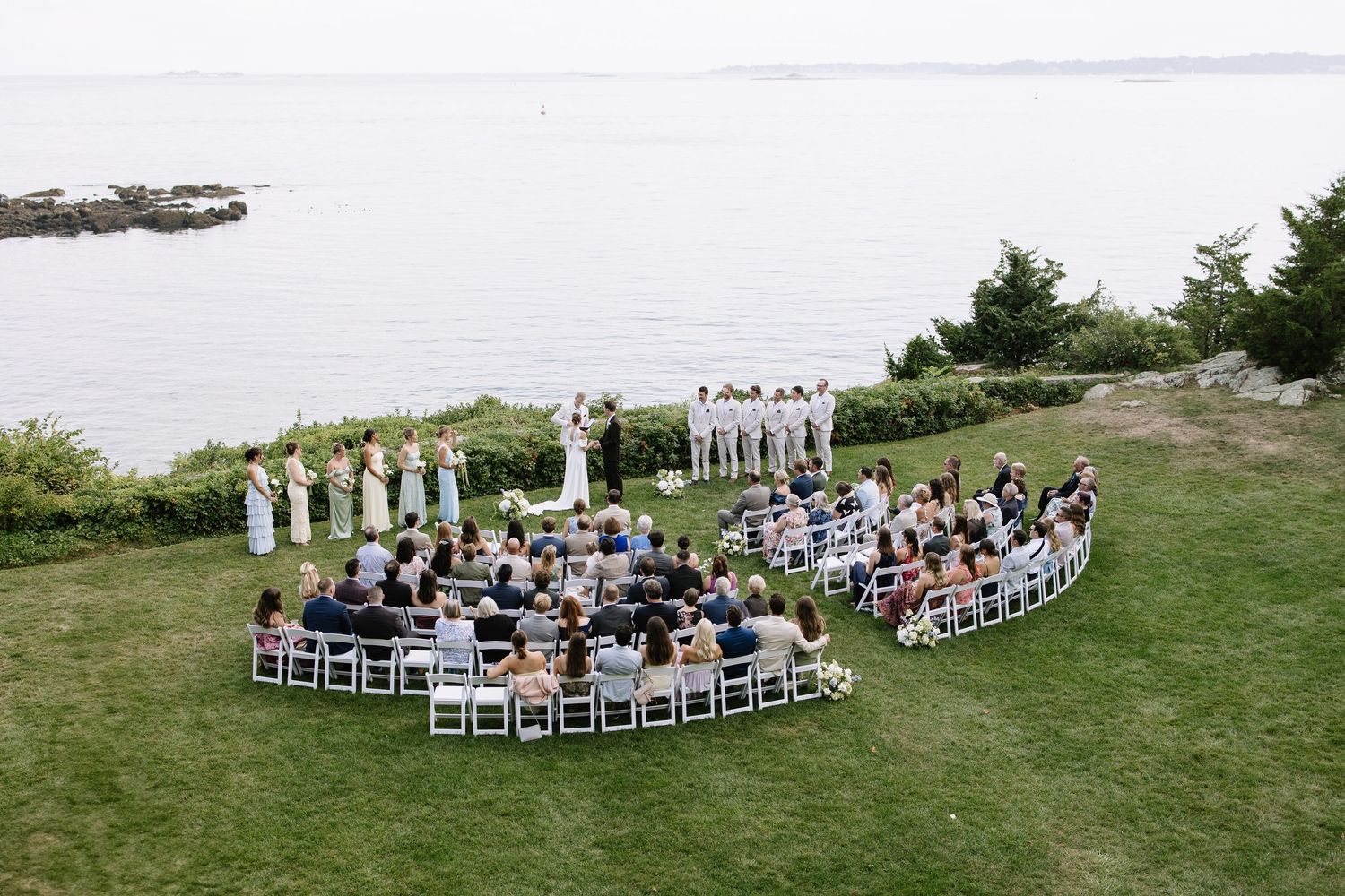 Outdoor wedding ceremony on a grassy cliff overlooking the ocean, with guests seated in a semicircle of white chairs.