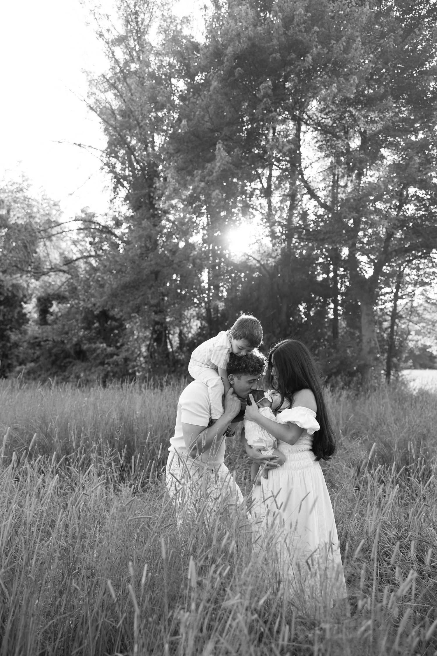 Black and white photo of a family with young child sharing a tender moment in a sunlit meadow with tall grass and trees.