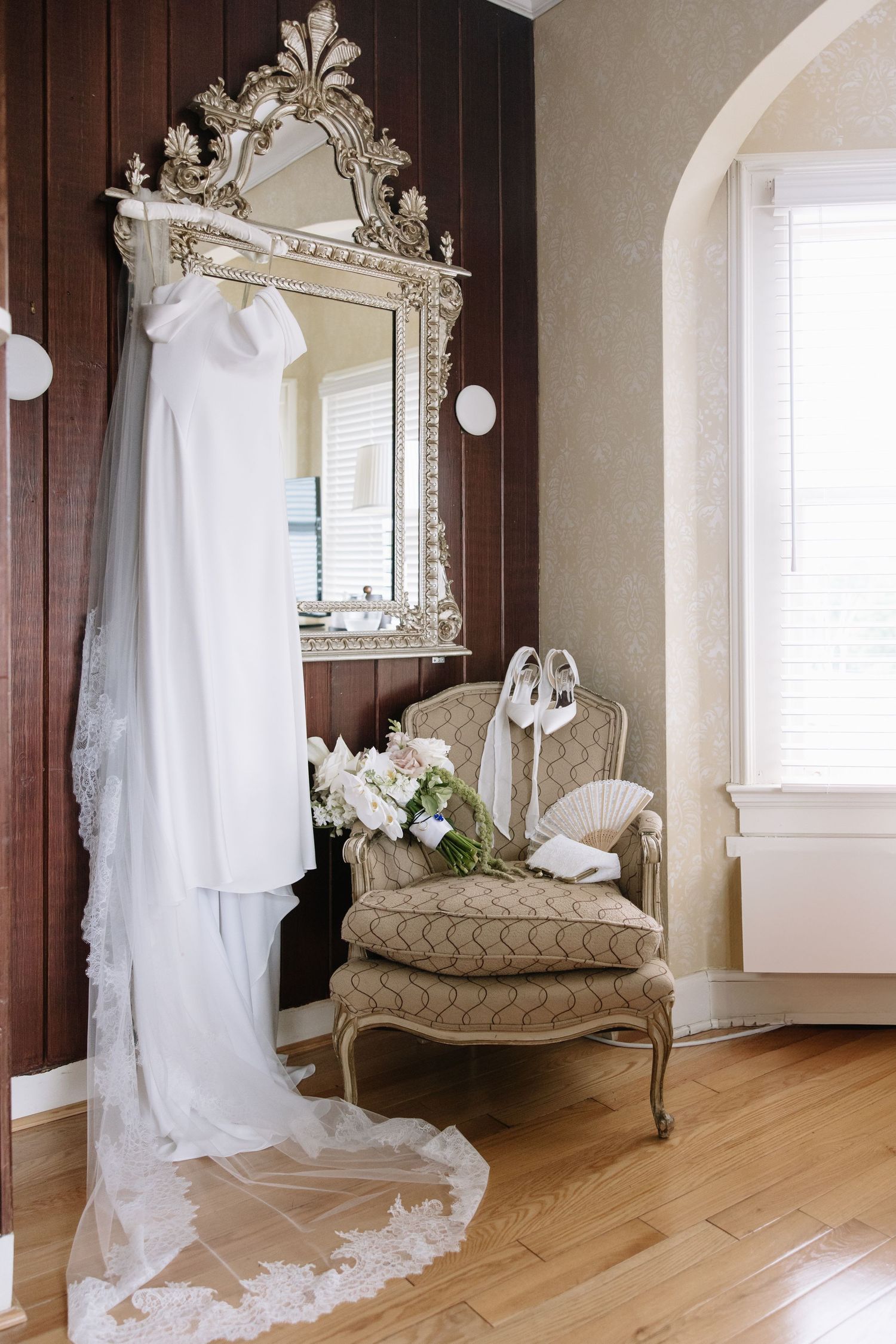 Elegant wedding dress hanging on ornate silver mirror beside vintage French chair with bridal bouquet and shoes.