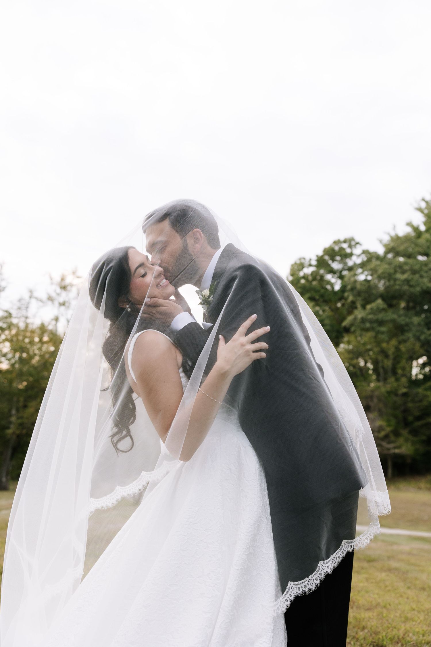 Bride and groom share a romantic kiss under a flowing white veil outdoors on their wedding day.