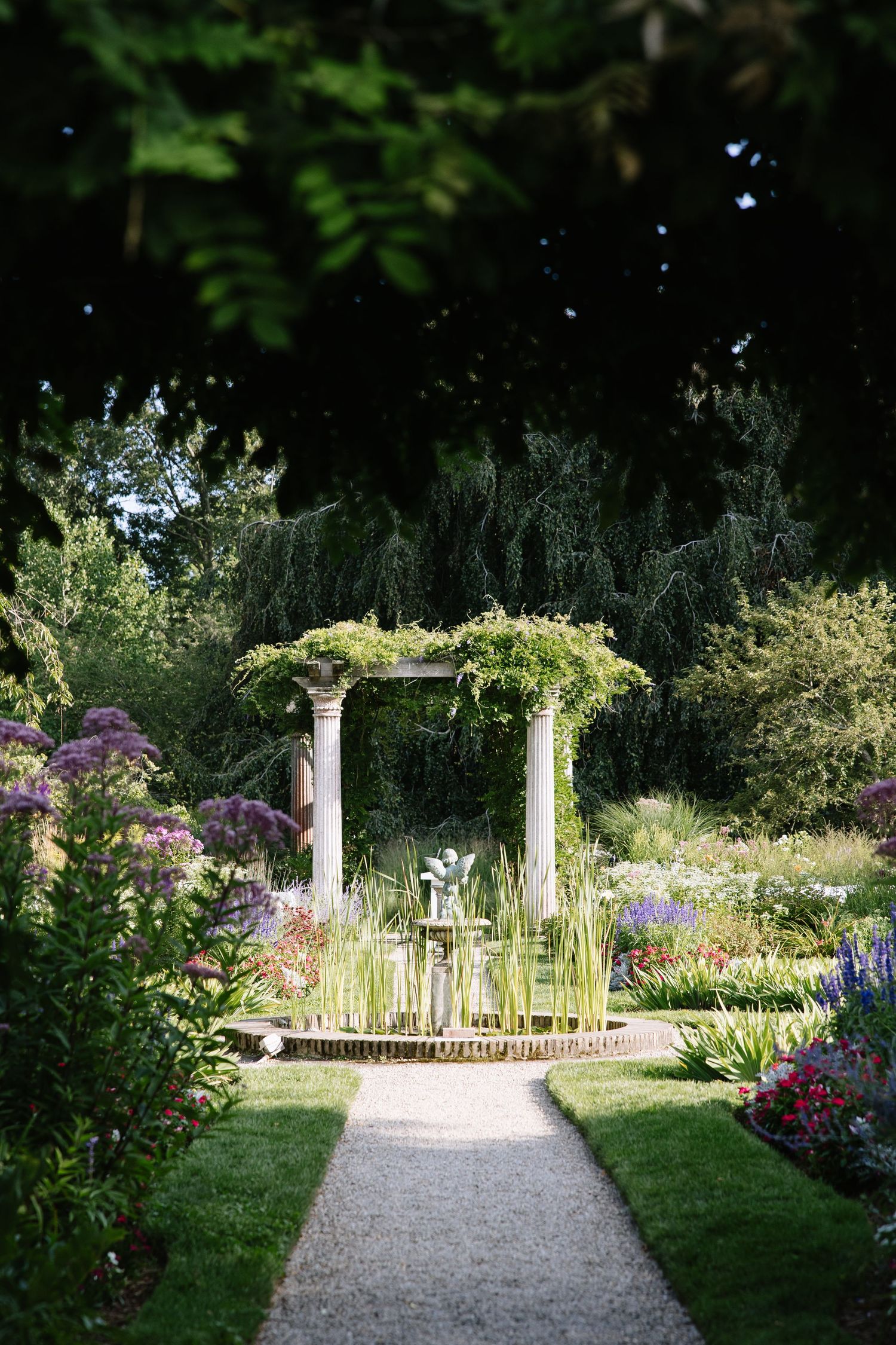 Elegant garden pergola with stone columns draped in greenery, surrounded by colorful flowering plants and a gravel path.