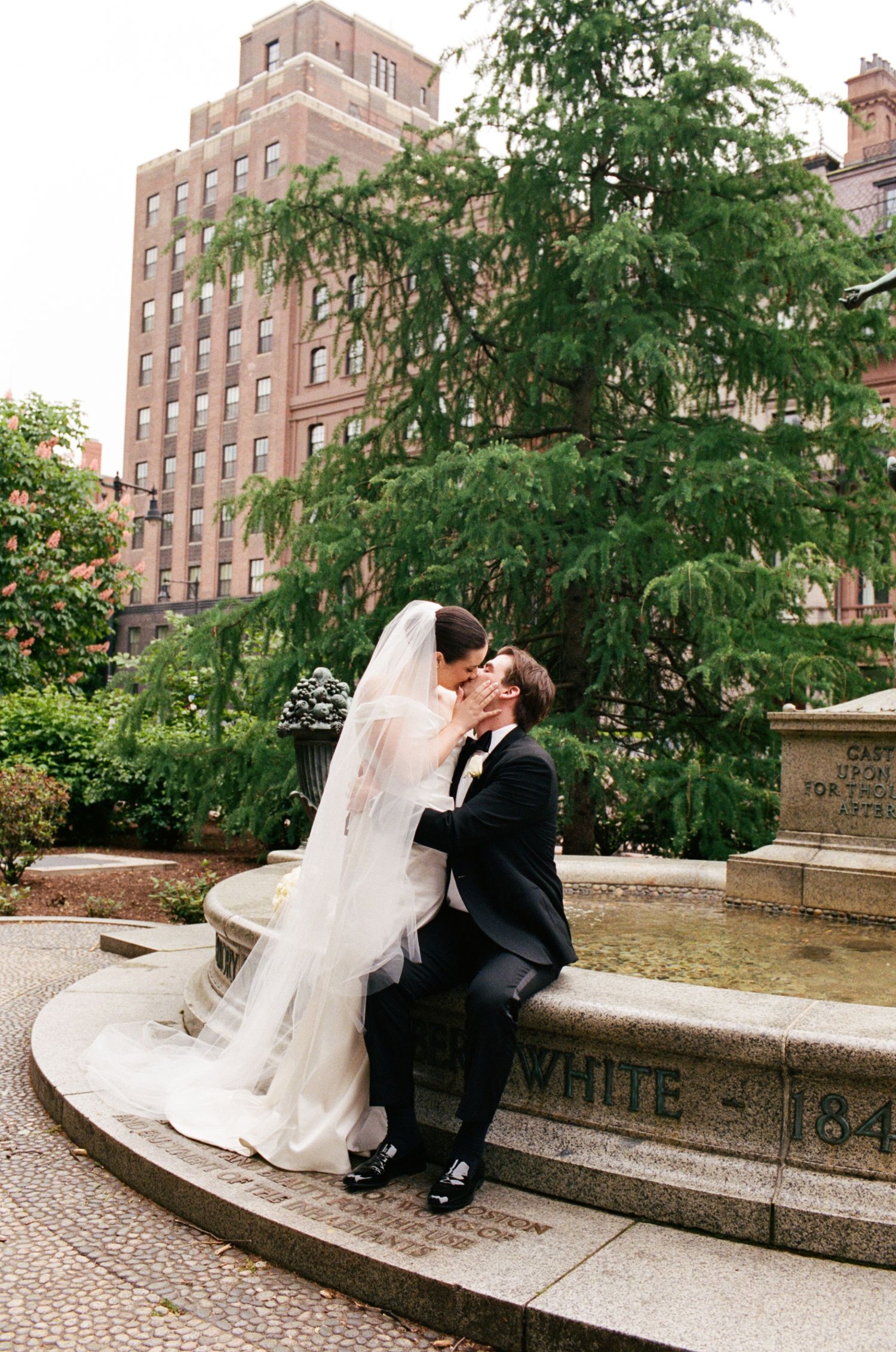 A bride and groom share a romantic kiss while seated at an outdoor fountain, surrounded by lush greenery and a classic brick building.
