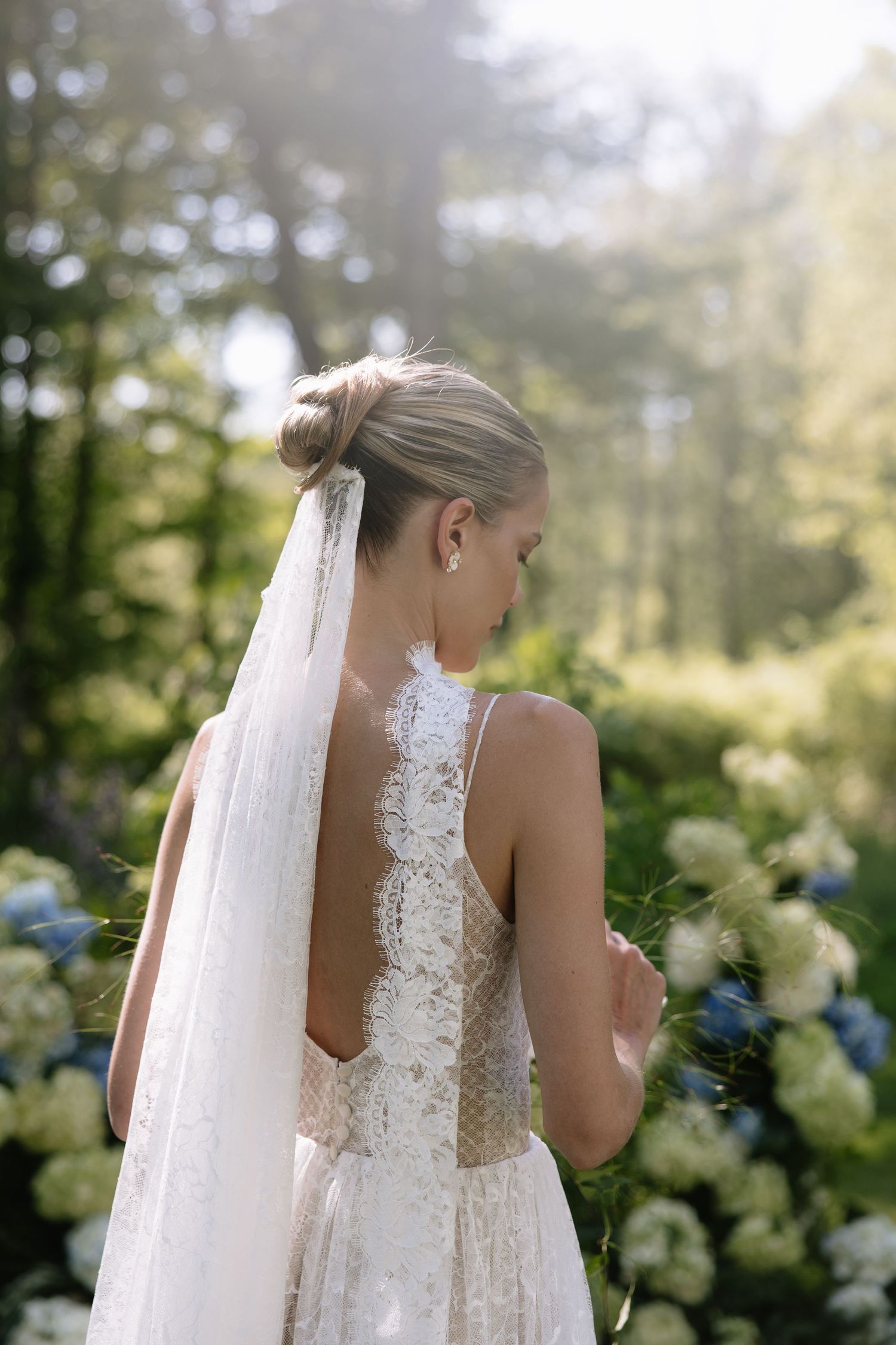 Bride in lace backless wedding dress with veil stands among hydrangeas in sunlit garden.