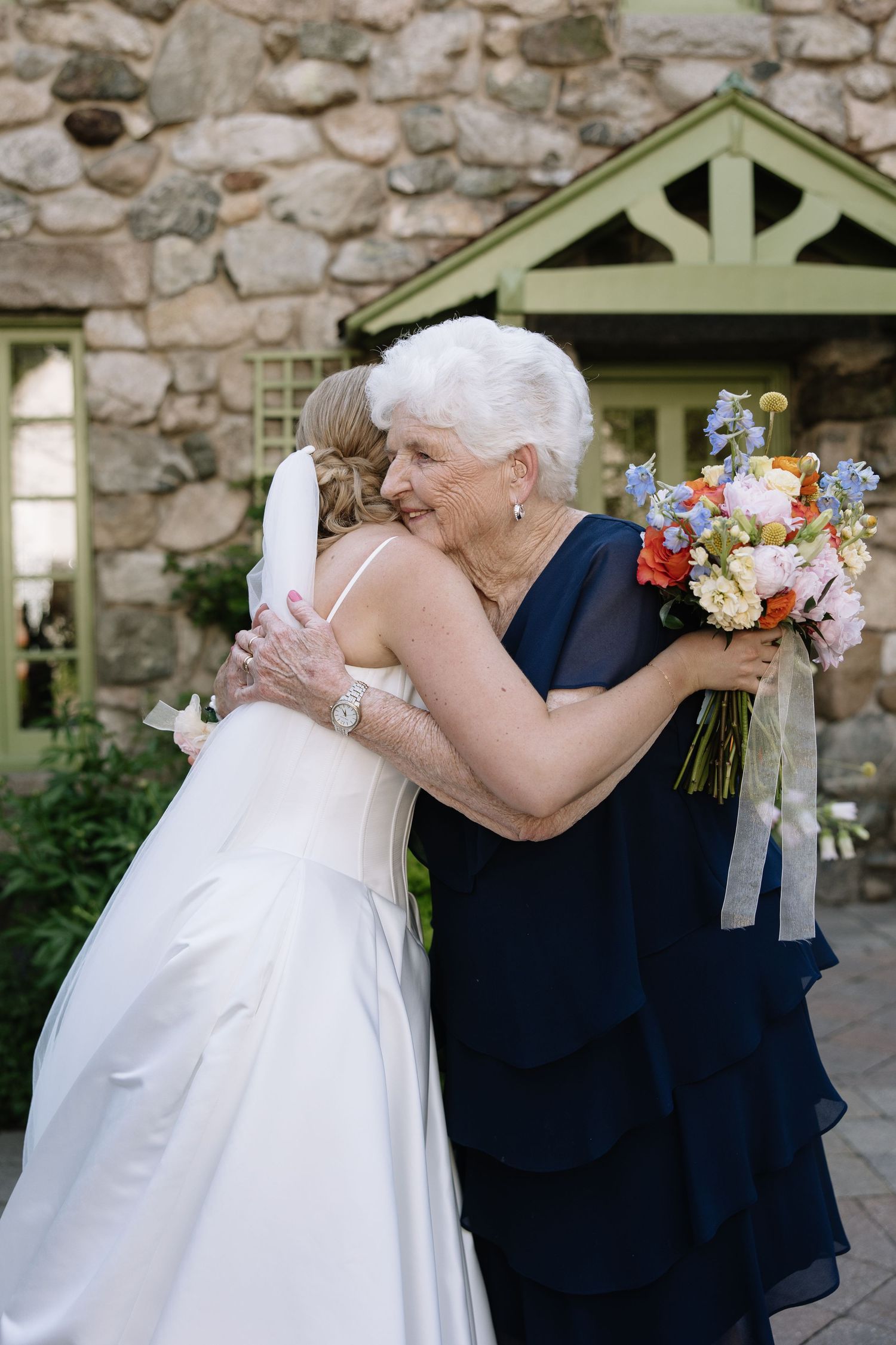 Bride in white gown hugging elderly woman in navy dress holding colorful bouquet outside stone building.