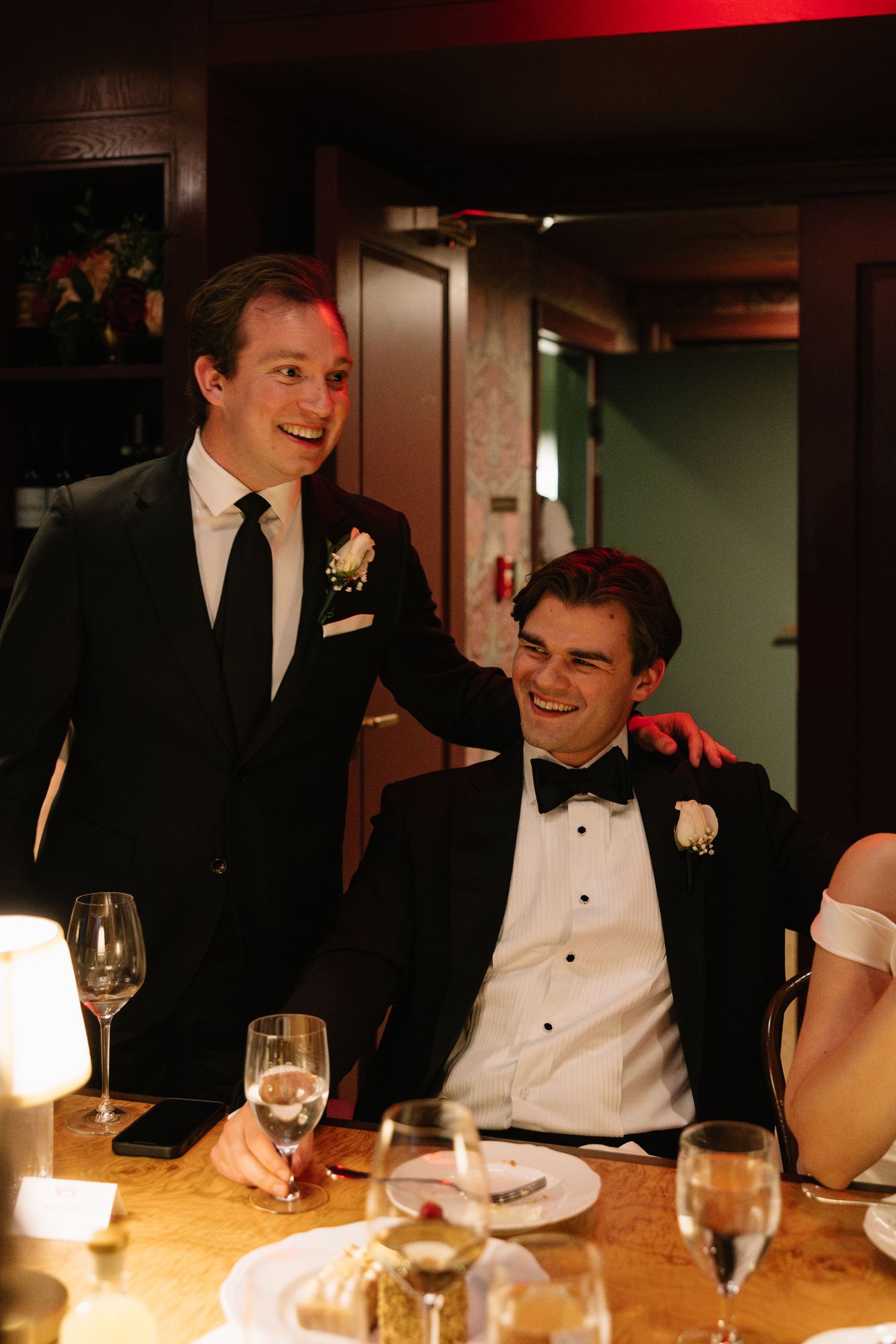 Two men in formal tuxedos smiling at a wedding reception dinner table with wine glasses.