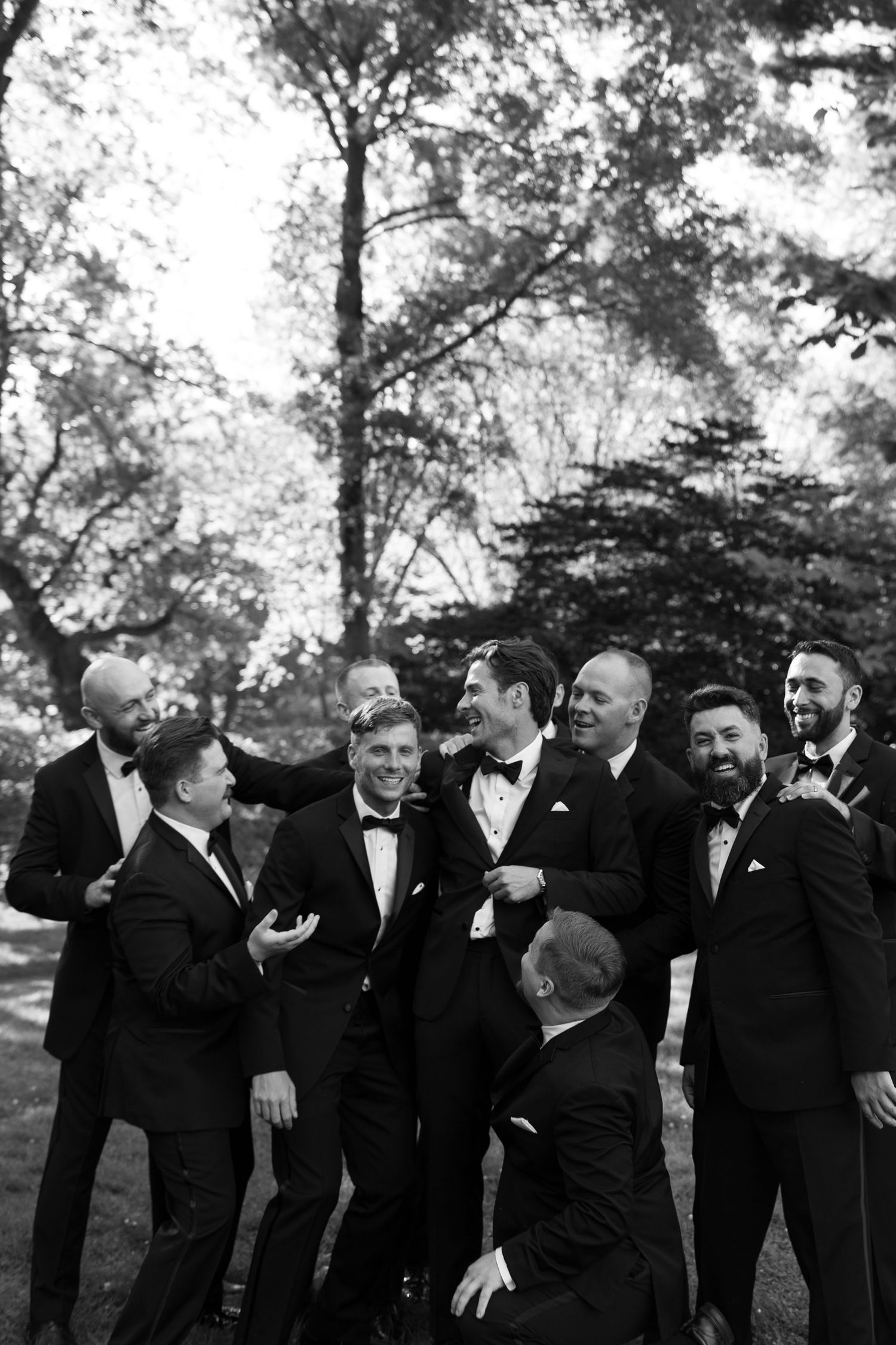 Black and white photo of a groom laughing with groomsmen in tuxedos outdoors at a wedding celebration.