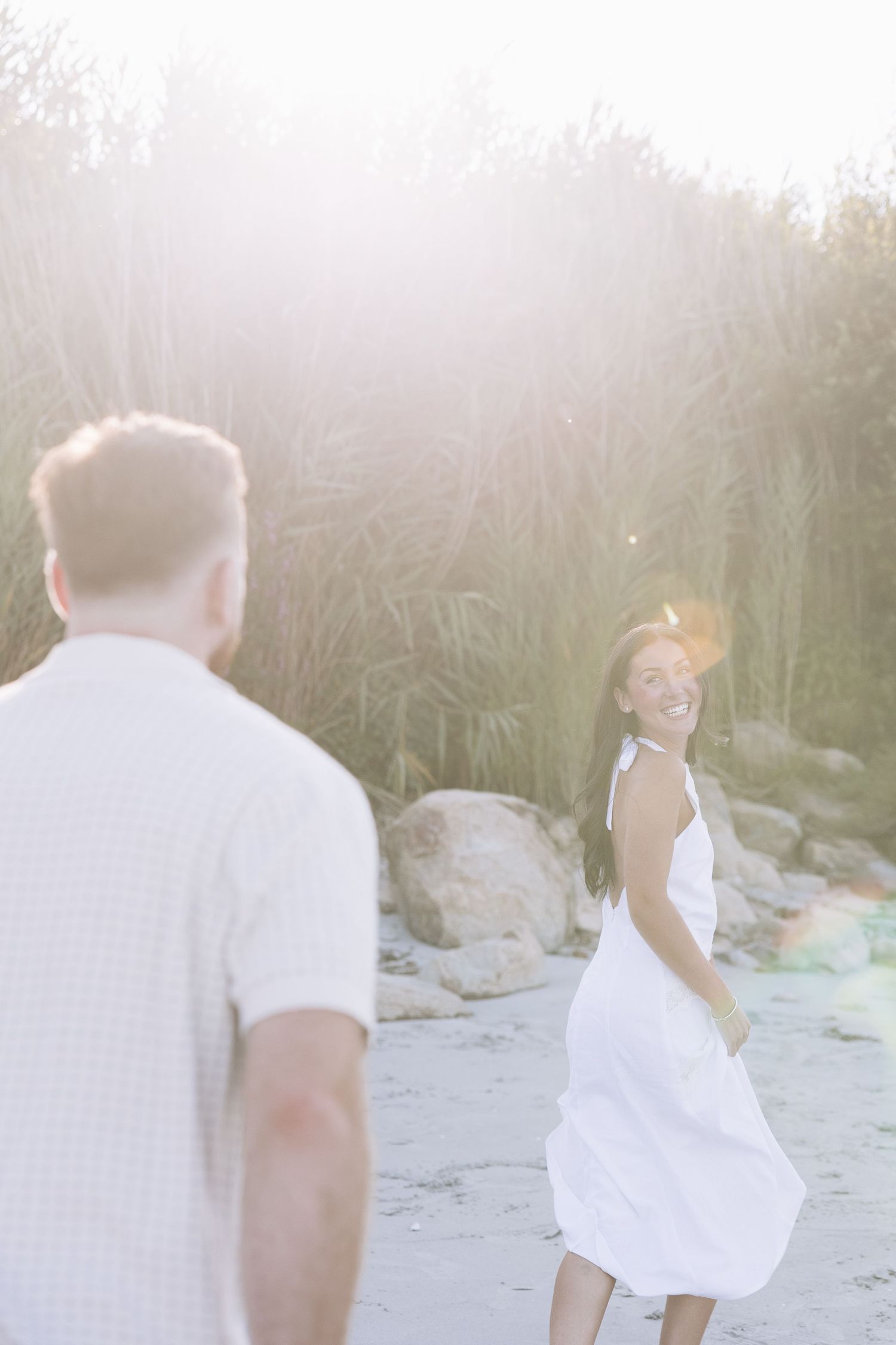 A smiling woman in a white dress looks back at a man during a golden hour outdoor photo session near tall grasses.