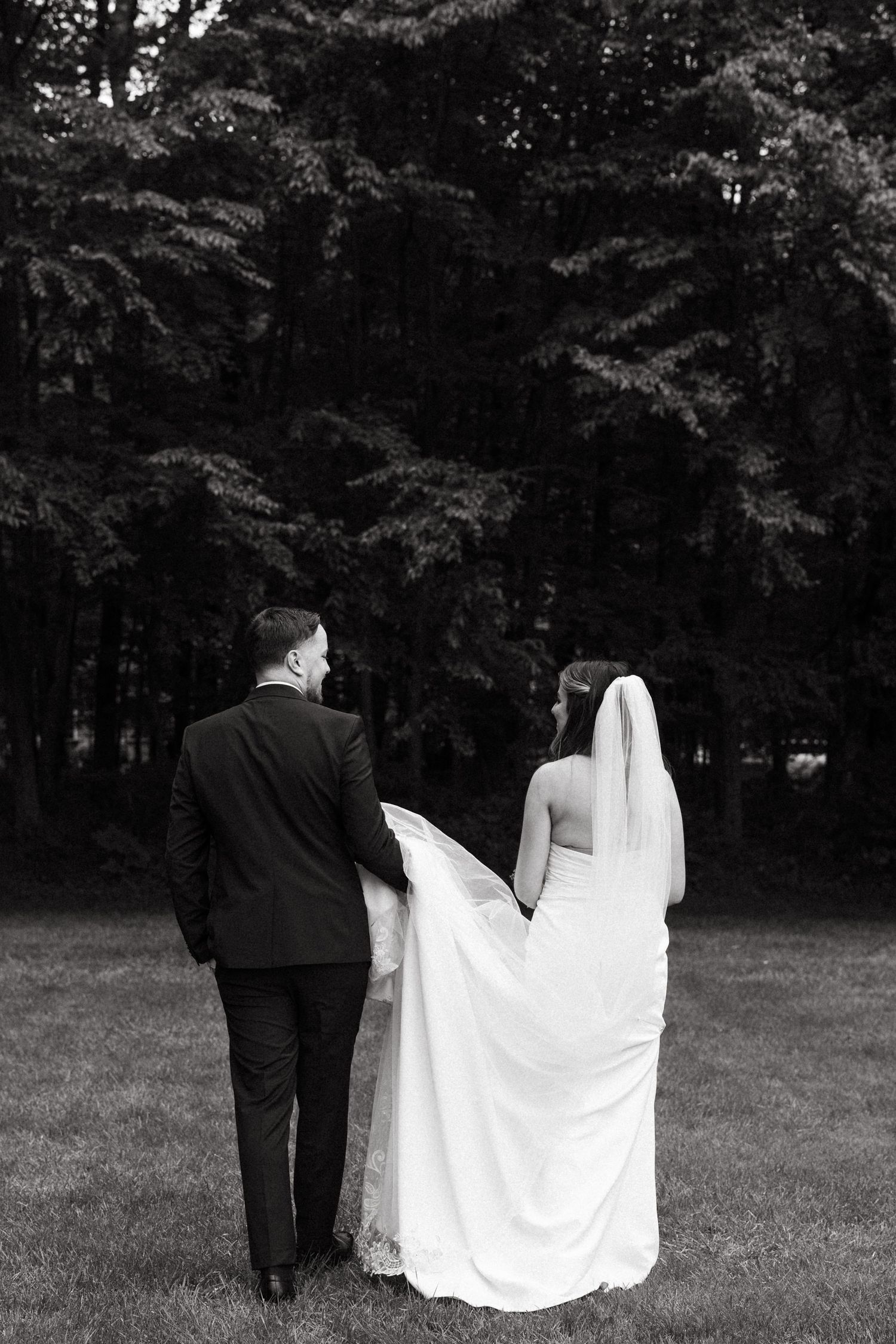 Couple walks away from camera across grass with bride's white dress trailing behind.