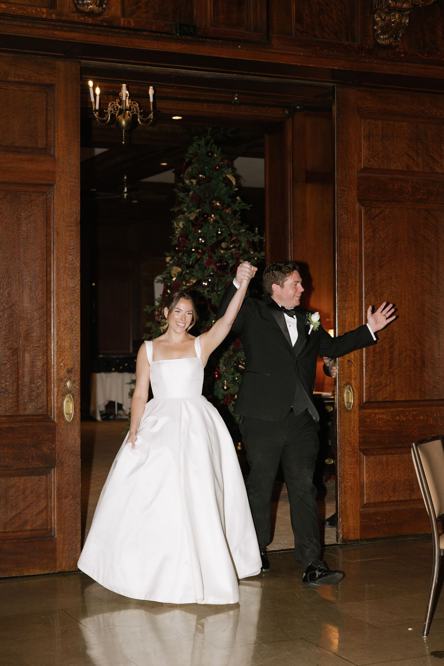 Wedding couple celebrates with joyful dance moves in wood-paneled ballroom.