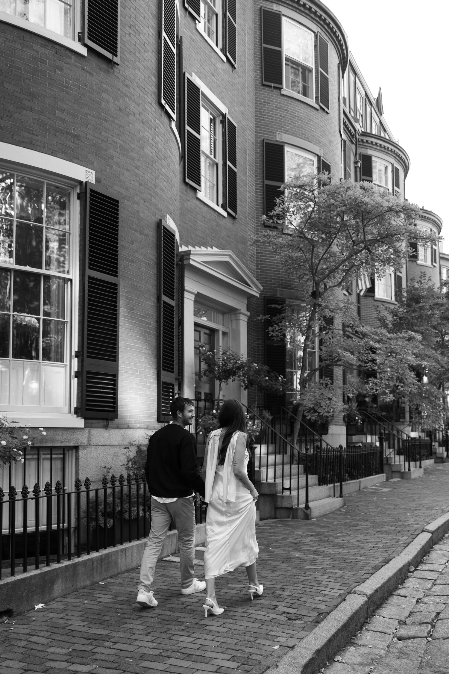 Black and white photo of couple walking along historic cobblestone street with brick buildings.