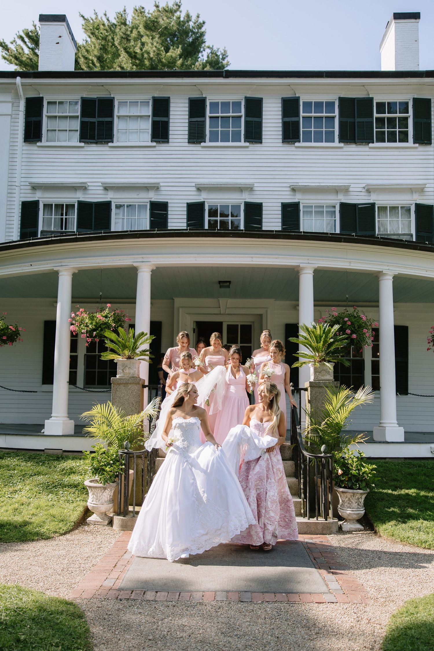 Wedding party in pink dresses poses on white columned mansion porch with flowers and greenery.