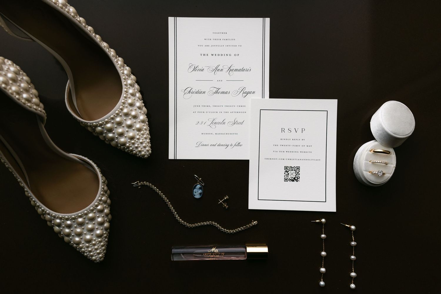 Wedding invitation suite with pearl-studded bridal shoes and jewelry on black background.