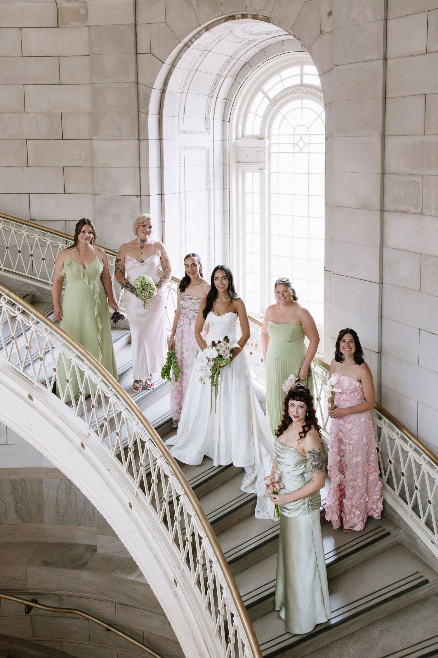 Bride in white gown poses with bridesmaids in sage green and pink dresses on elegant marble staircase.