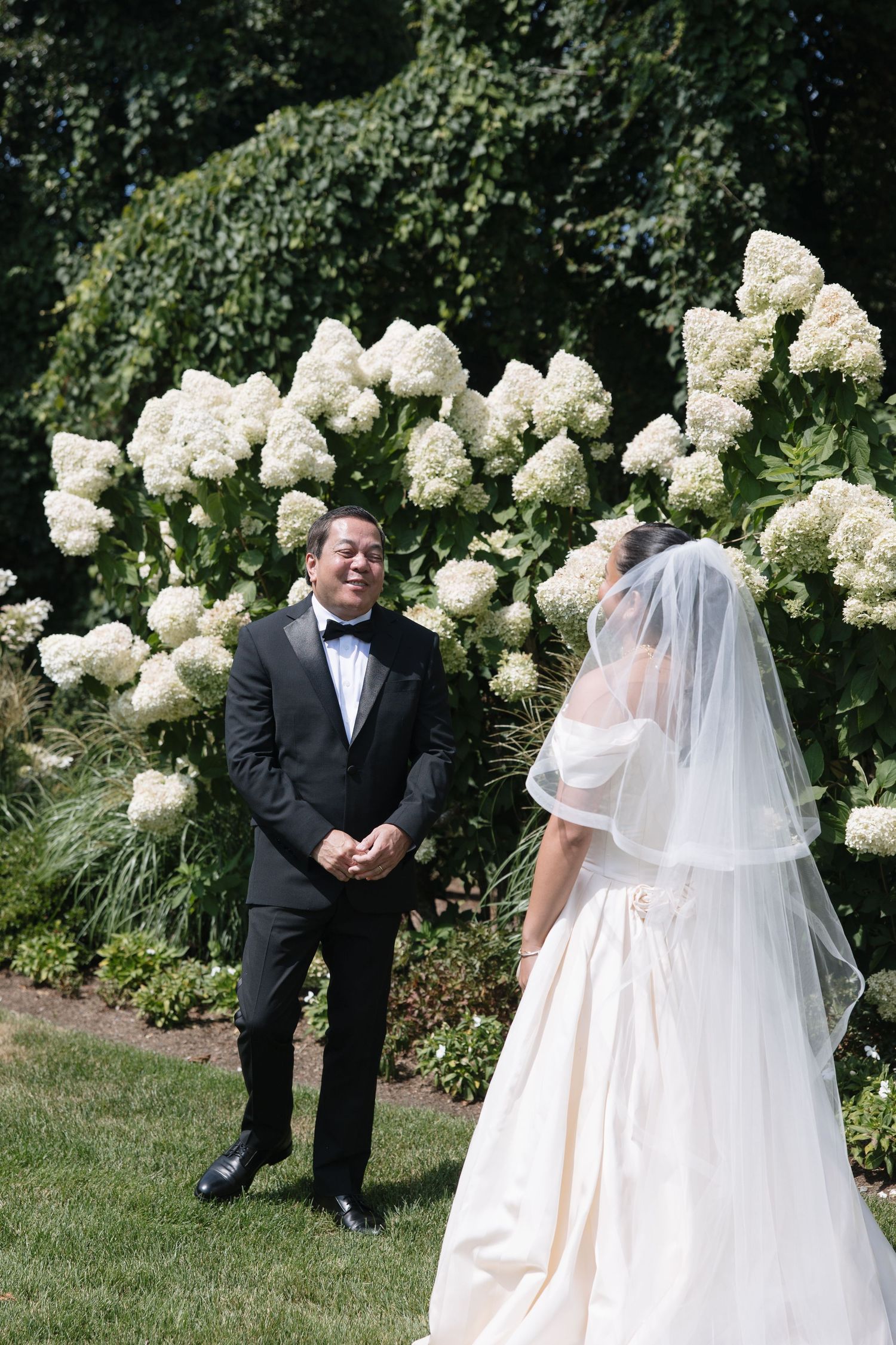 Groom sees bride for first time in front of white hydrangea bushes.