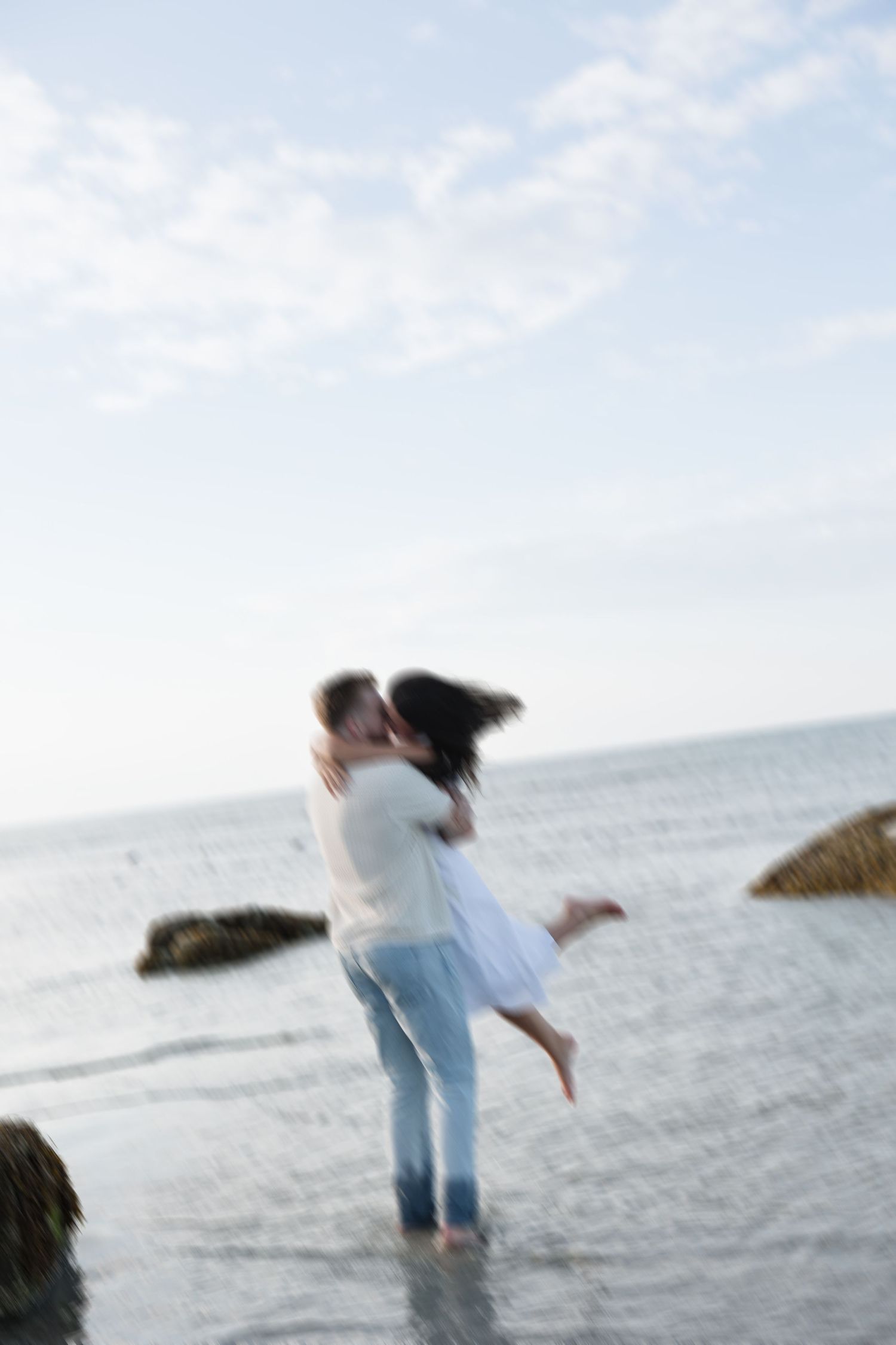 Couple sharing a romantic kiss by the ocean, man lifting woman in white dress near rocky shoreline.