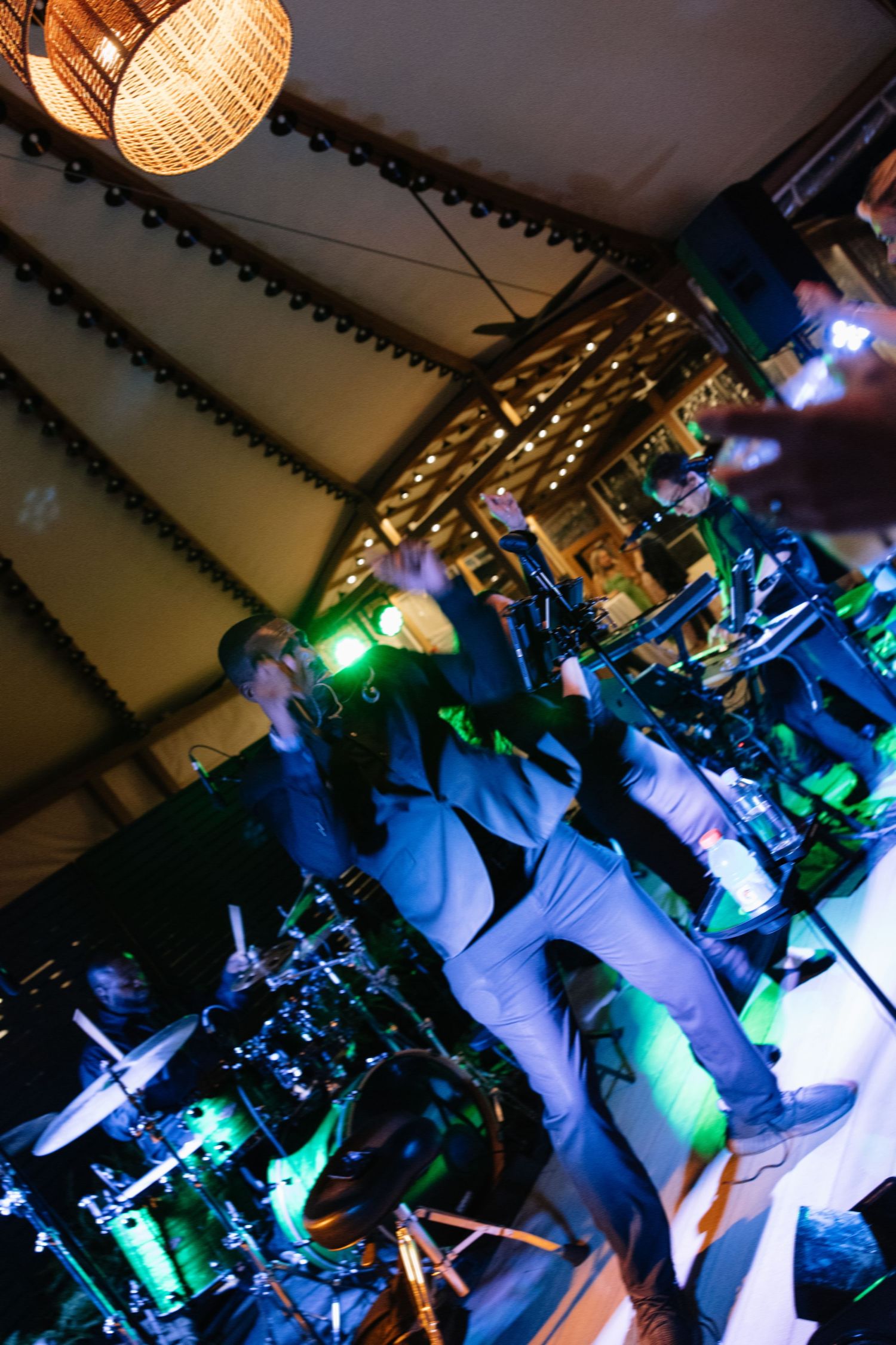 Live band performing energetically under tent venue lights with drums and green stage lighting creating dynamic motion blur effect.