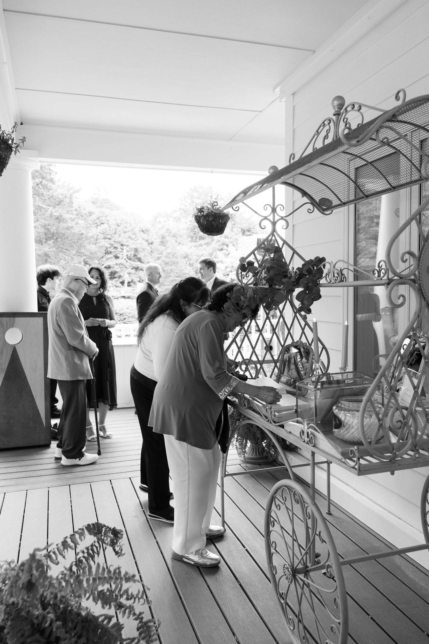Black and white photo of people browsing an ornate metal display cart on a wooden porch at an outdoor event.