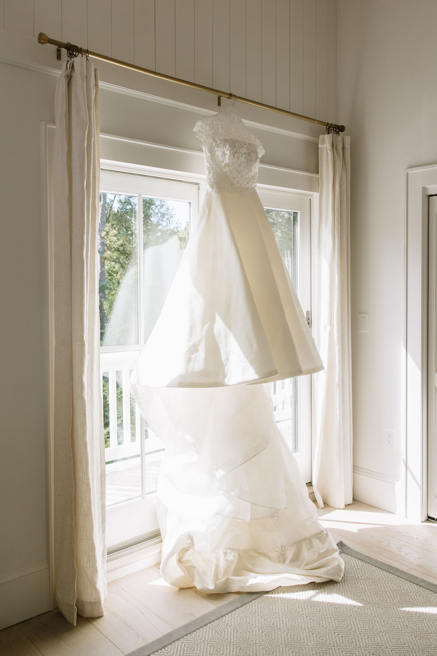 White wedding dress hanging in sunlit window creates ethereal silhouette.