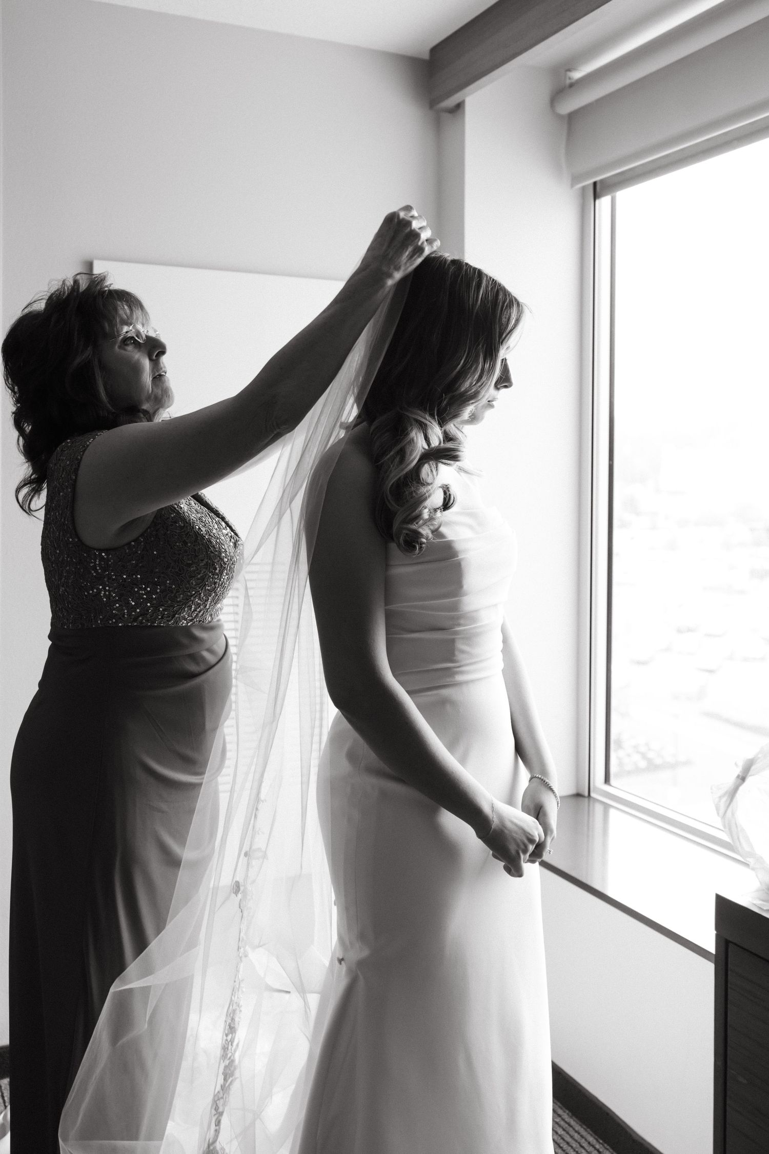 Intimate moment of wedding veil adjustment by window in black and white photography.