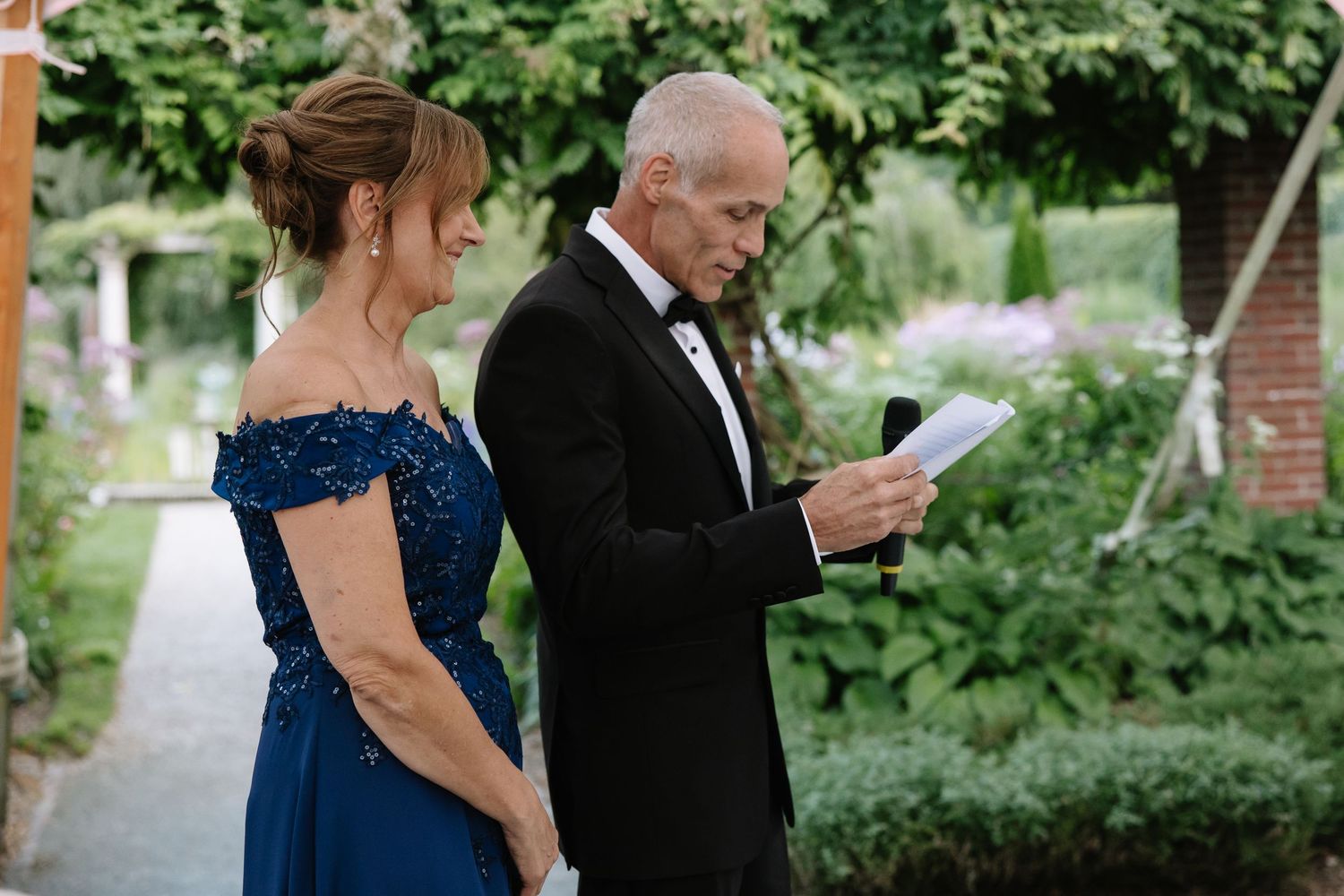 Wedding guests in formal attire gather for speeches during an outdoor garden reception.