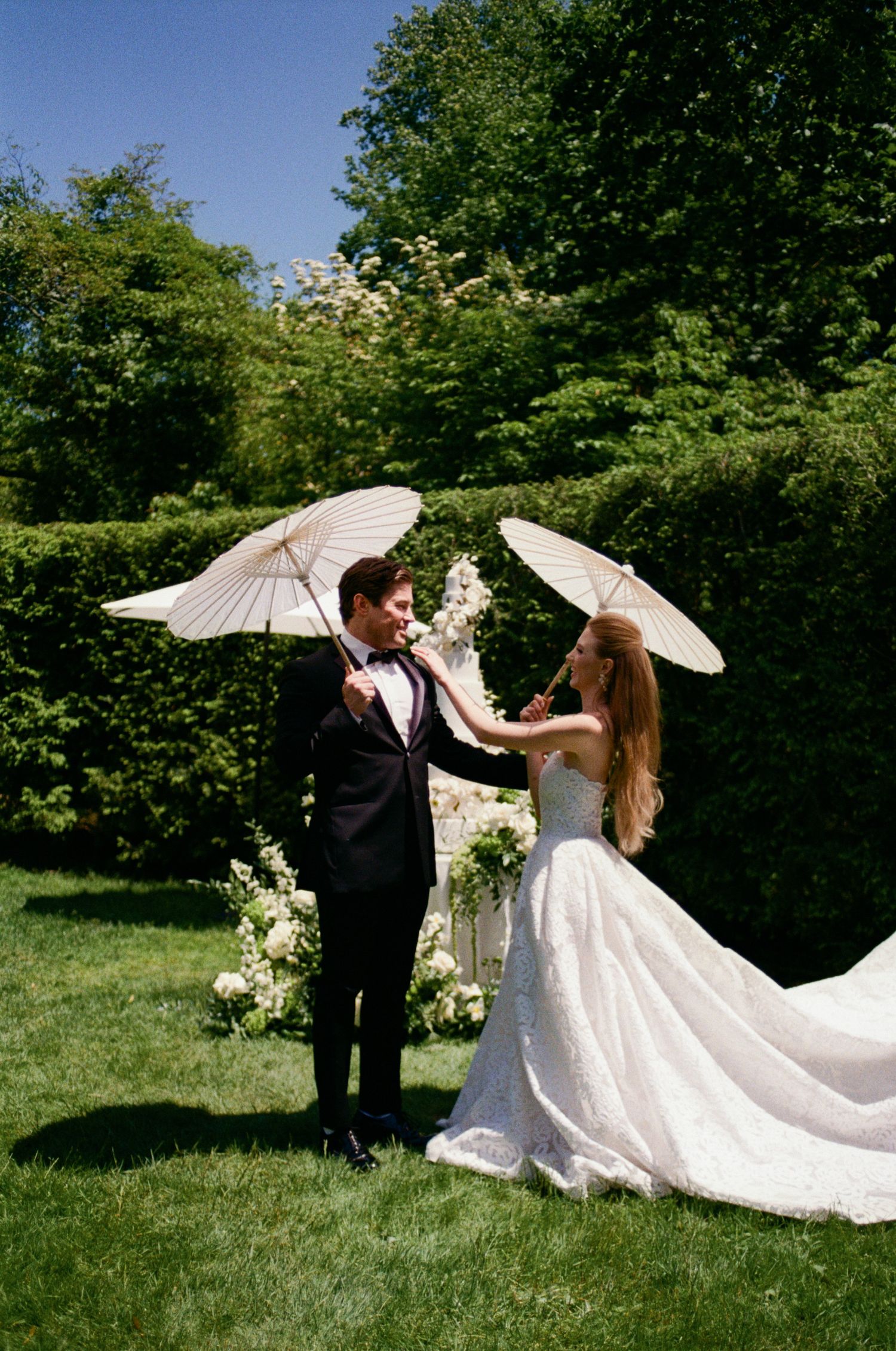 Couple with parasol posing in a sunlit garden with flowing white dress.