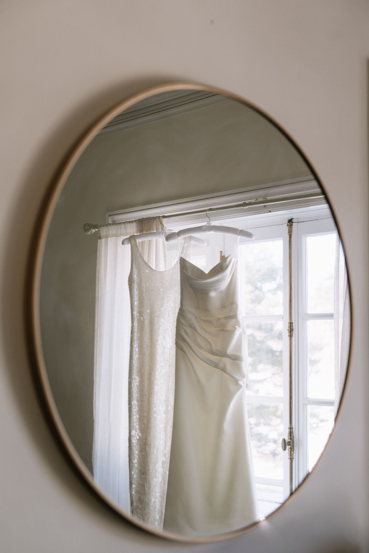 Wedding dress hanging in window reflection captured in round mirror on wall.