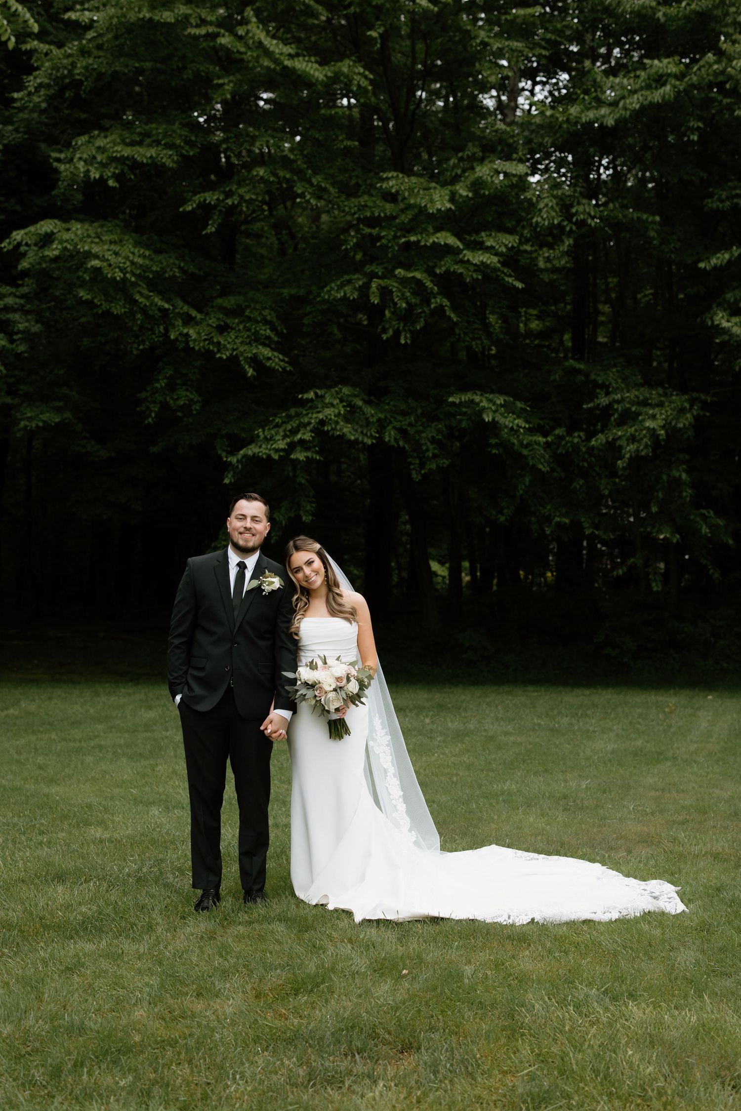 Bride in flowing white gown poses with bouquet on grassy lawn.