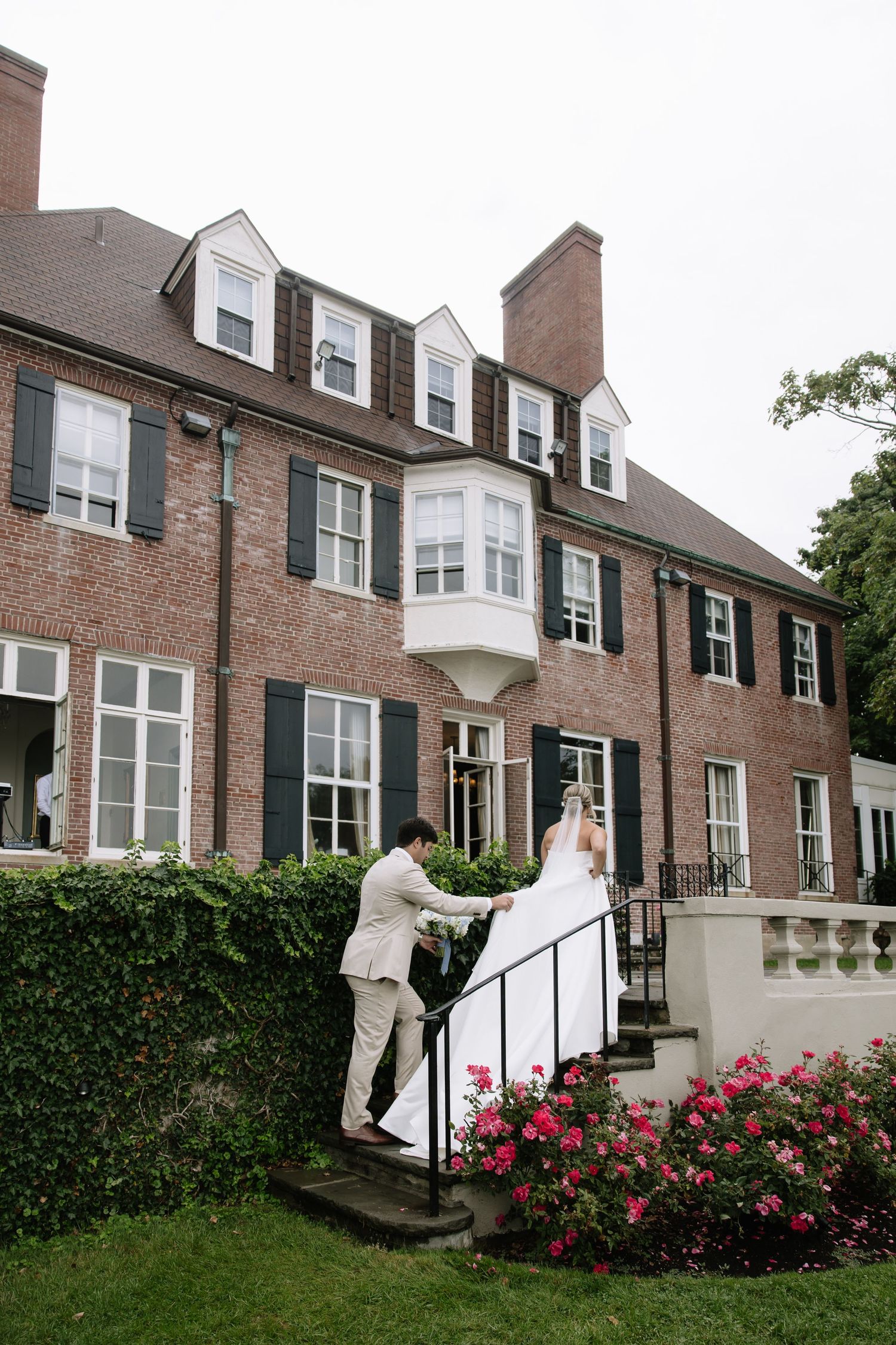 A couple ascends brick mansion steps surrounded by pink roses and landscaping.