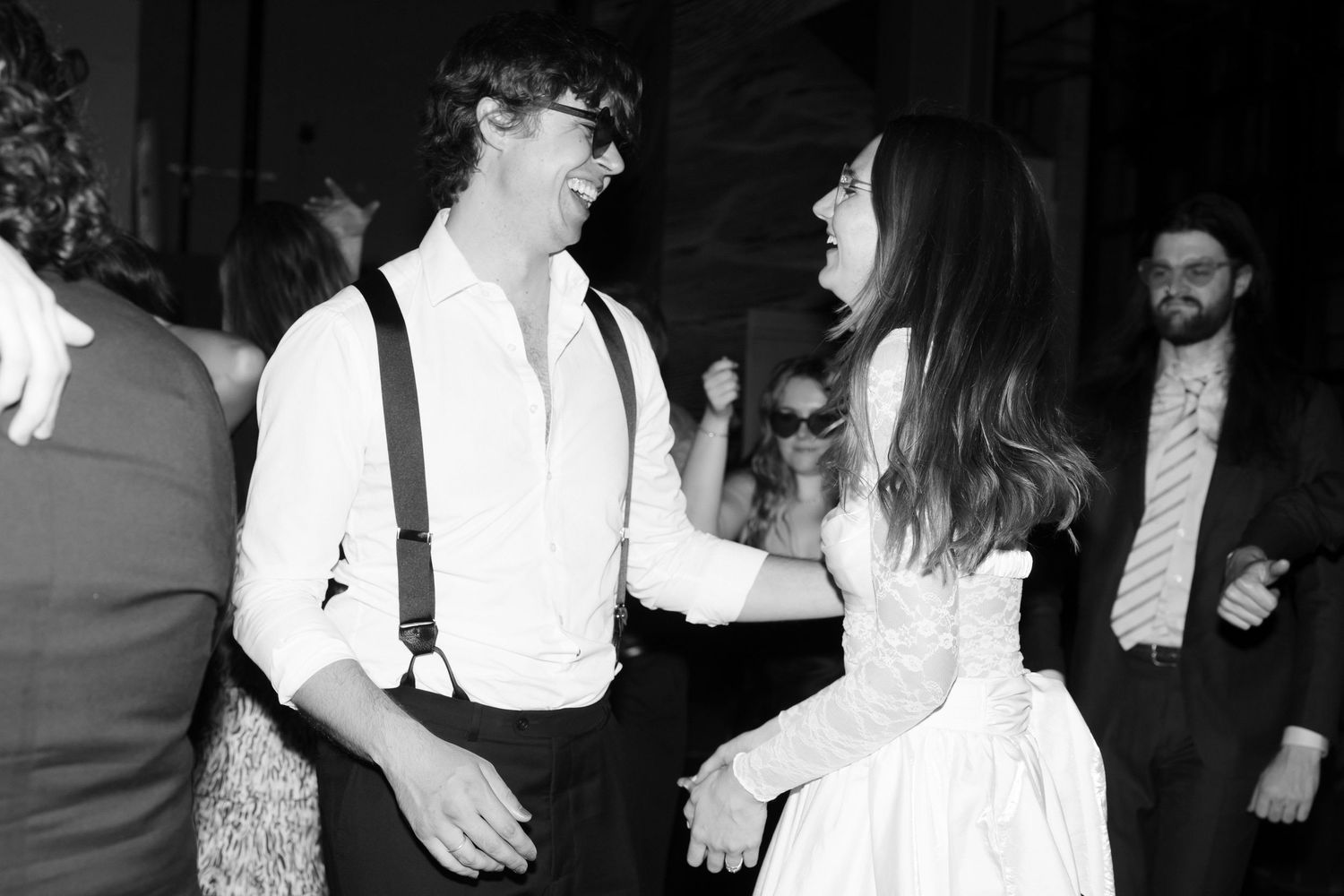 Black and white photo of a couple dancing joyfully at a wedding reception, man in suspenders and woman in white dress.