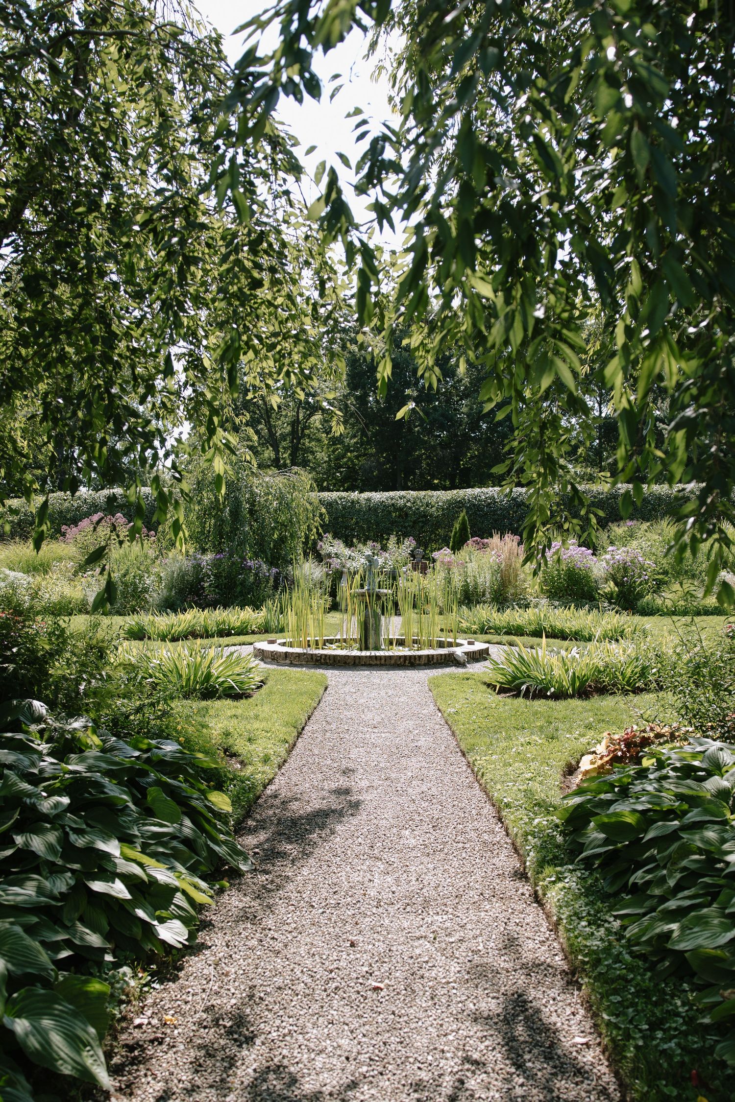 A serene garden path leads to a circular fountain surrounded by lush greenery and manicured hedges under a canopy of trees.