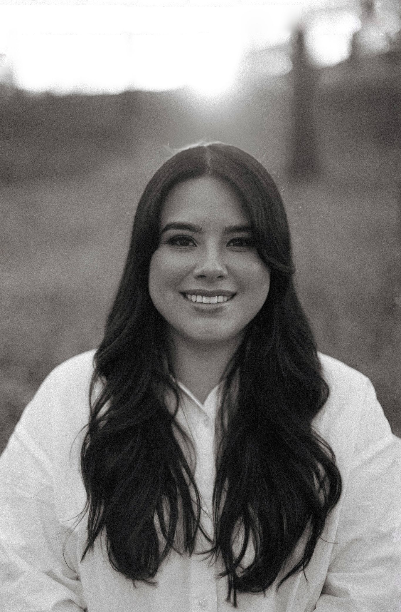Black and white portrait of a smiling young woman with long dark hair wearing a white shirt outdoors.