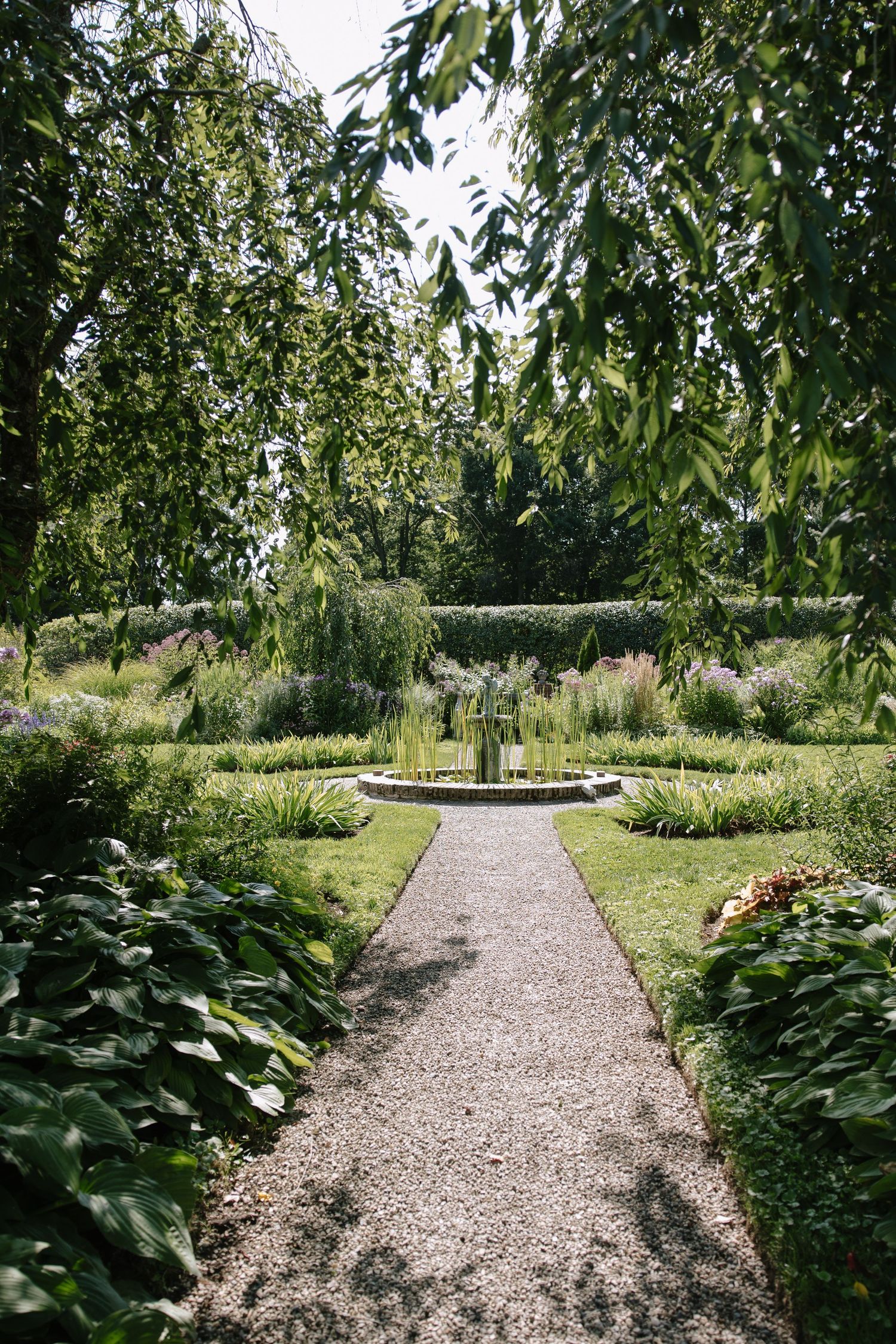 A serene garden path leads to a circular fountain surrounded by lush greenery and flowering plants beneath leafy trees.