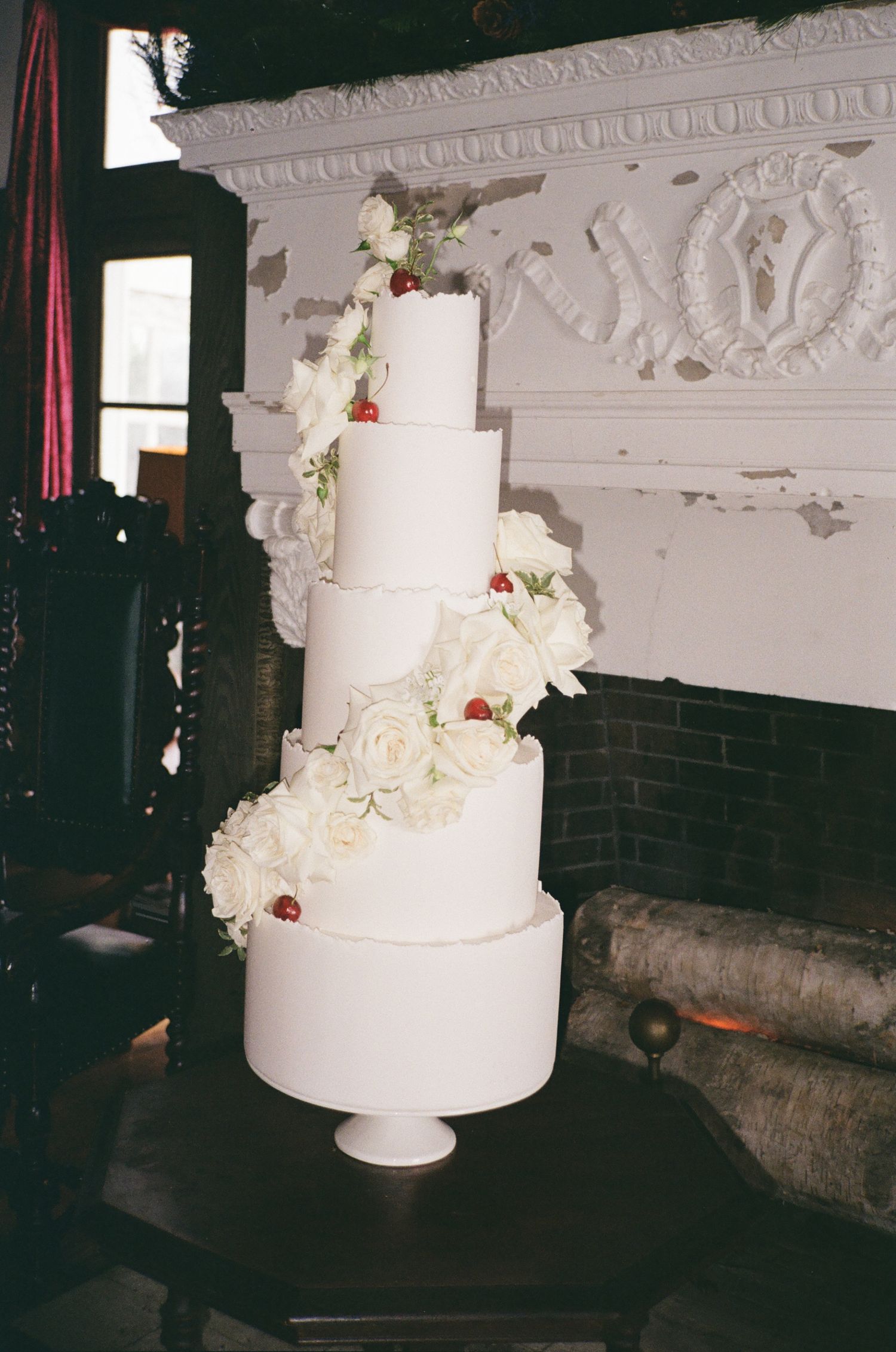 Elegant four-tier white wedding cake adorned with white roses and red berries, displayed near an ornate fireplace.