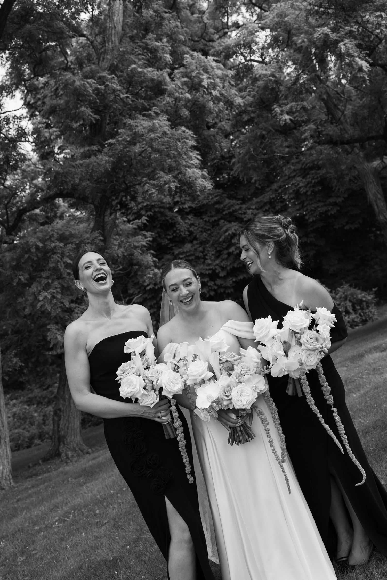 Black and white photo of a bride in white gown laughing with two bridesmaids in black dresses holding floral bouquets outdoors.