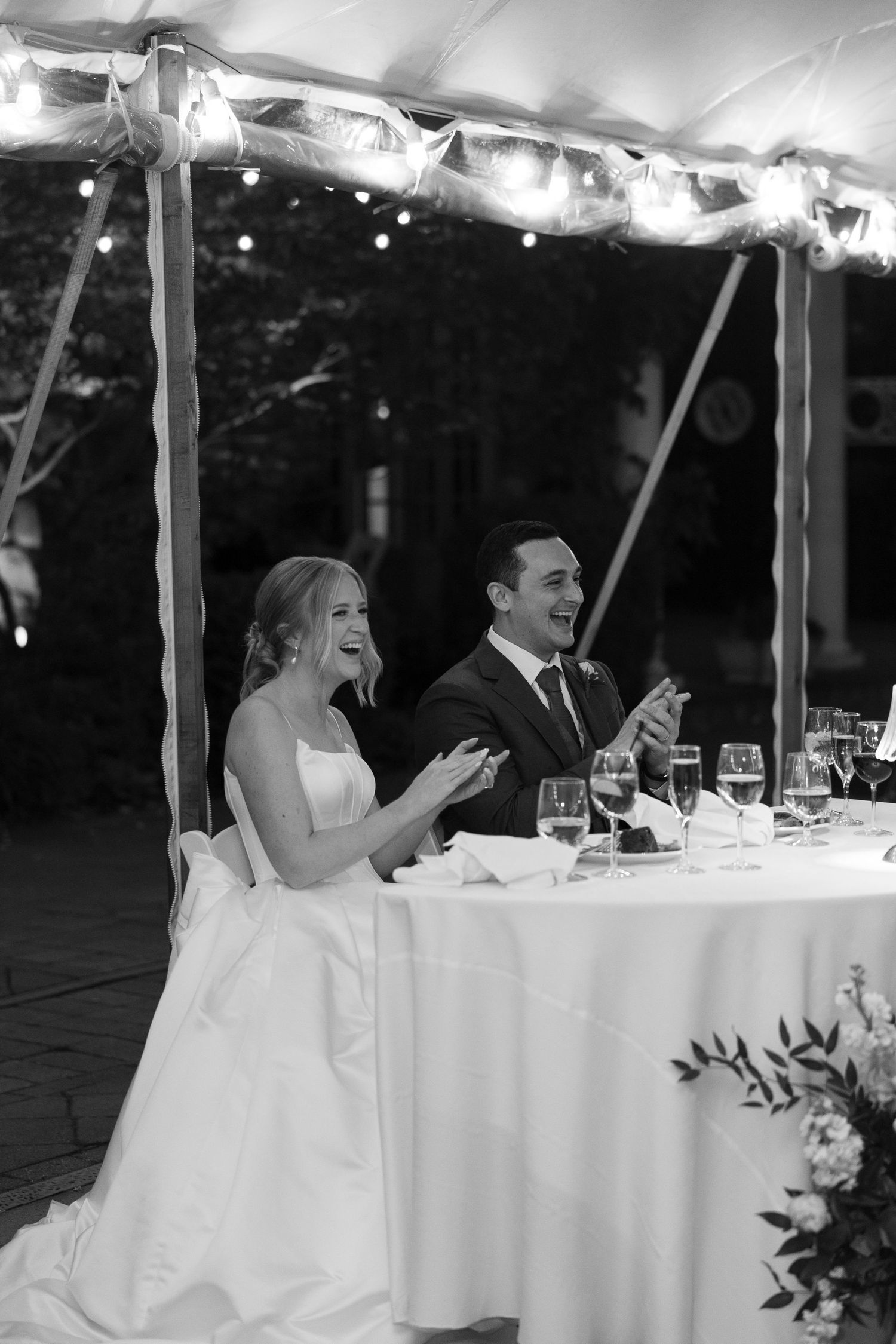 Bride and groom laugh and clap at their wedding reception table under a tent with string lights.